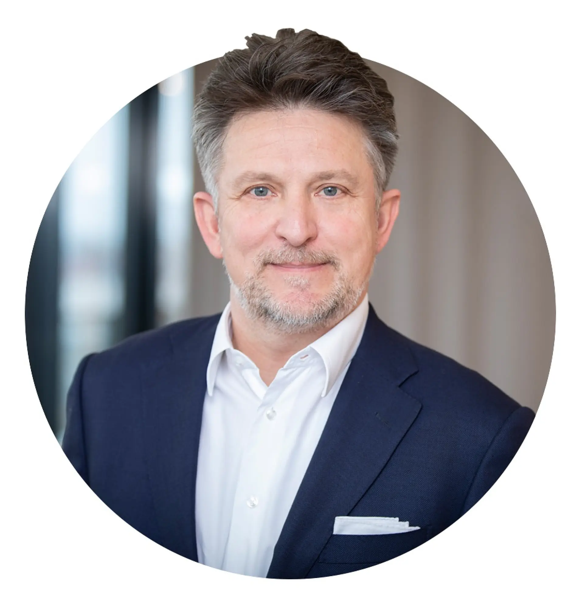 A man with short dark hair and a beard is wearing a navy blue suit, white shirt, and pocket square. He is smiling slightly while standing in front of a blurred background. The image is cropped in a circular format. (This text has been generated by AI)