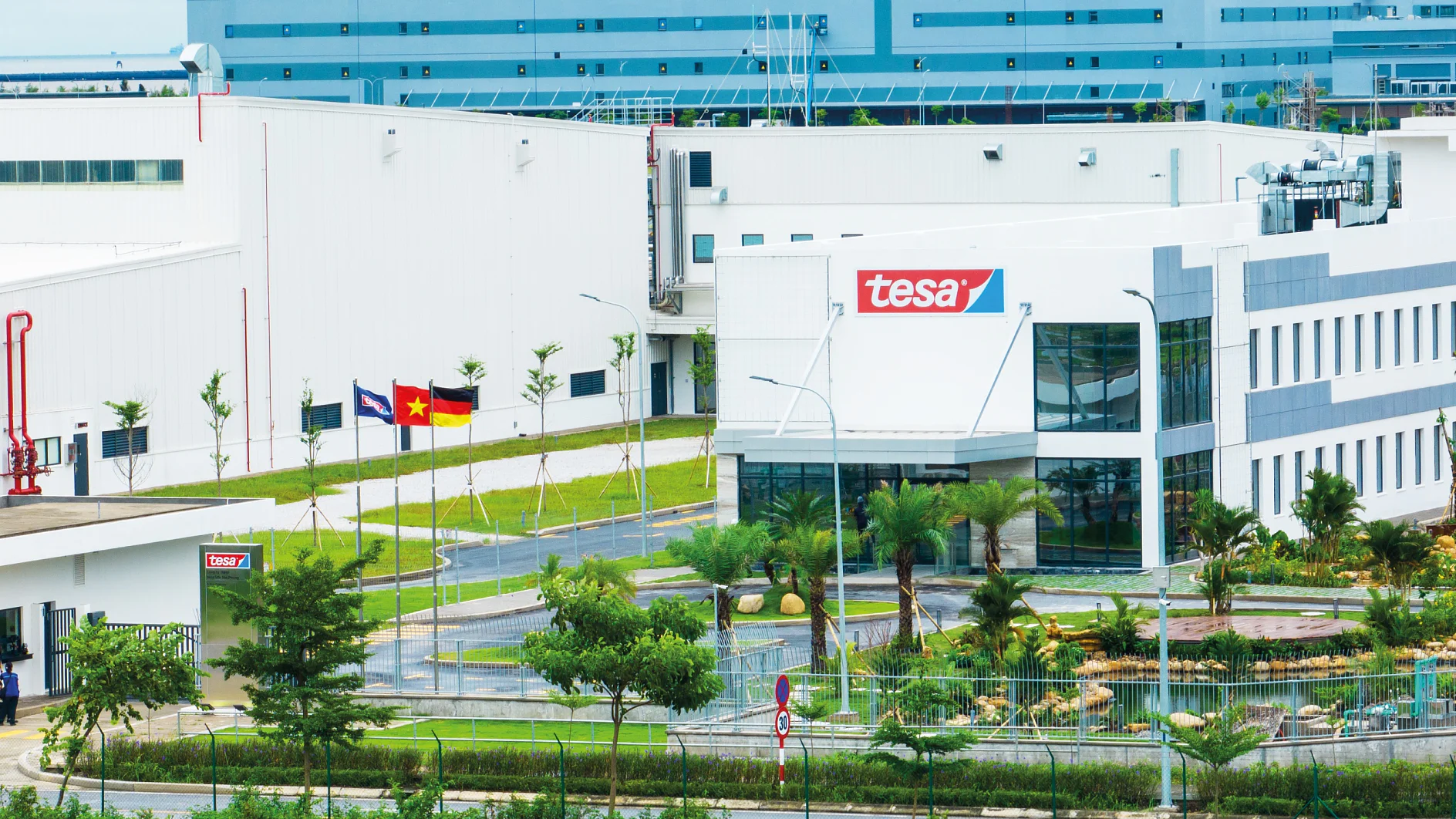 Exterior view of a modern industrial building with the logo tesa on it. The facility is surrounded by landscaped greenery, positioned in front of larger blue and white buildings. Flags are visible near the entrance, showcasing their renowned product, tesa tape. (This text has been generated by AI)