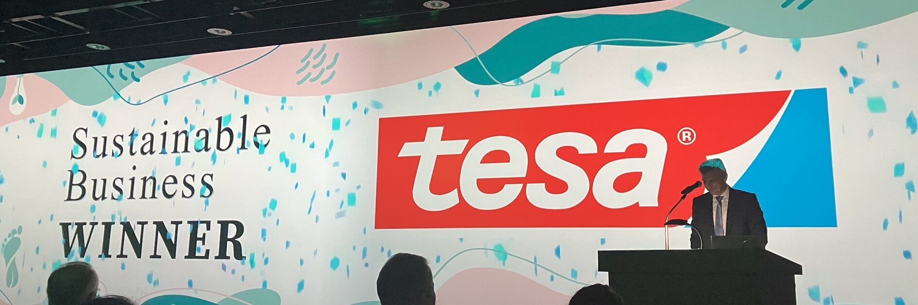 ステージ上の演壇に立つ人物の背後には、「tesa」という単語と「持続可能なビジネス ウィナー」というフレーズが表示された大きなスクリーンがあります。前景に観客が見えます。. (This text has been generated by AI)
