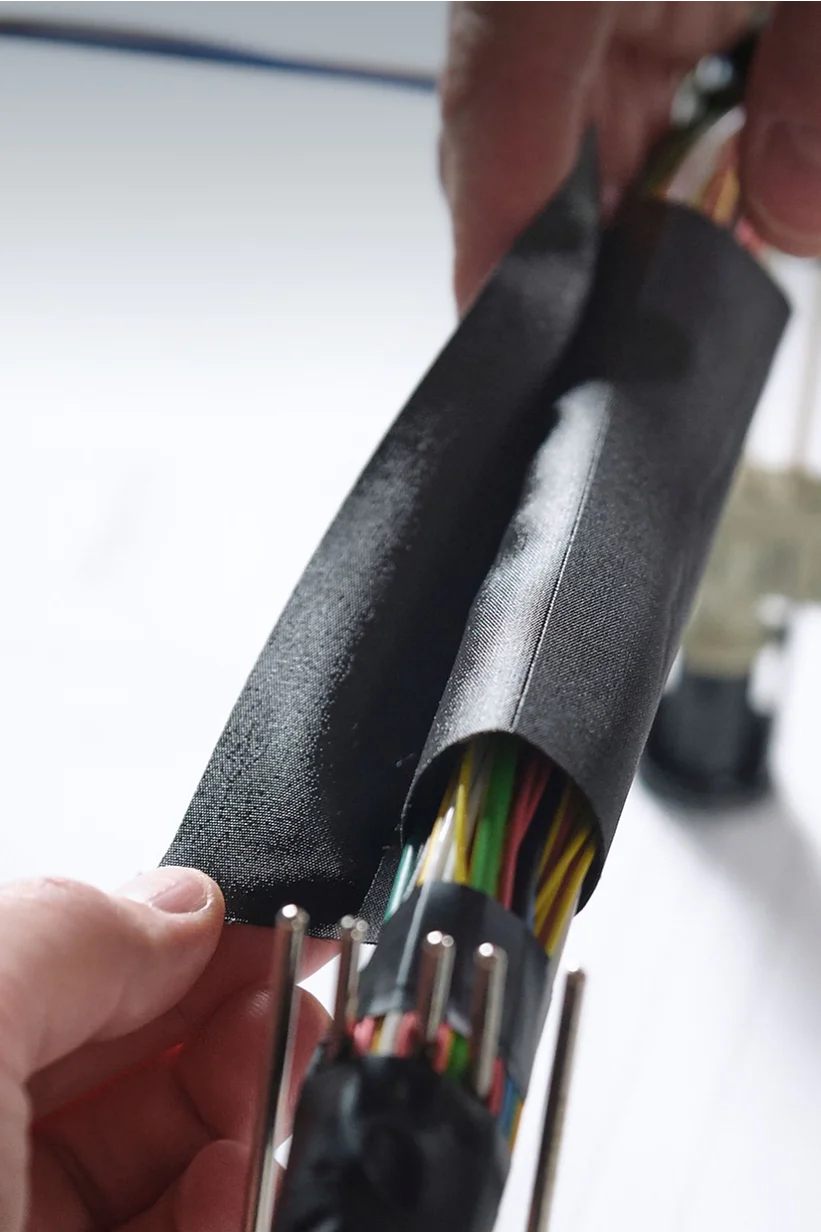 Close-up of hands wrapping a bundle of multicolored electrical wires with black insulating tesa tape. The wires are organized and connected to plastic connectors. A blurred background shows more wires and a surface with printed text. (This text has been generated by AI)