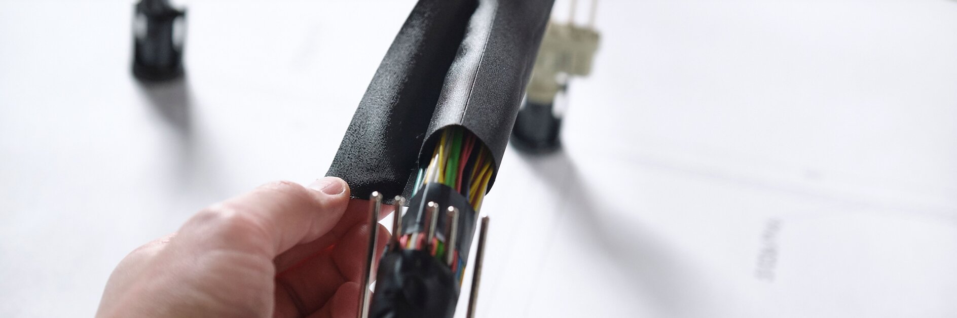 Close-up of hands wrapping a bundle of multicolored electrical wires with black insulating tesa tape. The wires are organized and connected to plastic connectors. A blurred background shows more wires and a surface with printed text. (This text has been generated by AI)