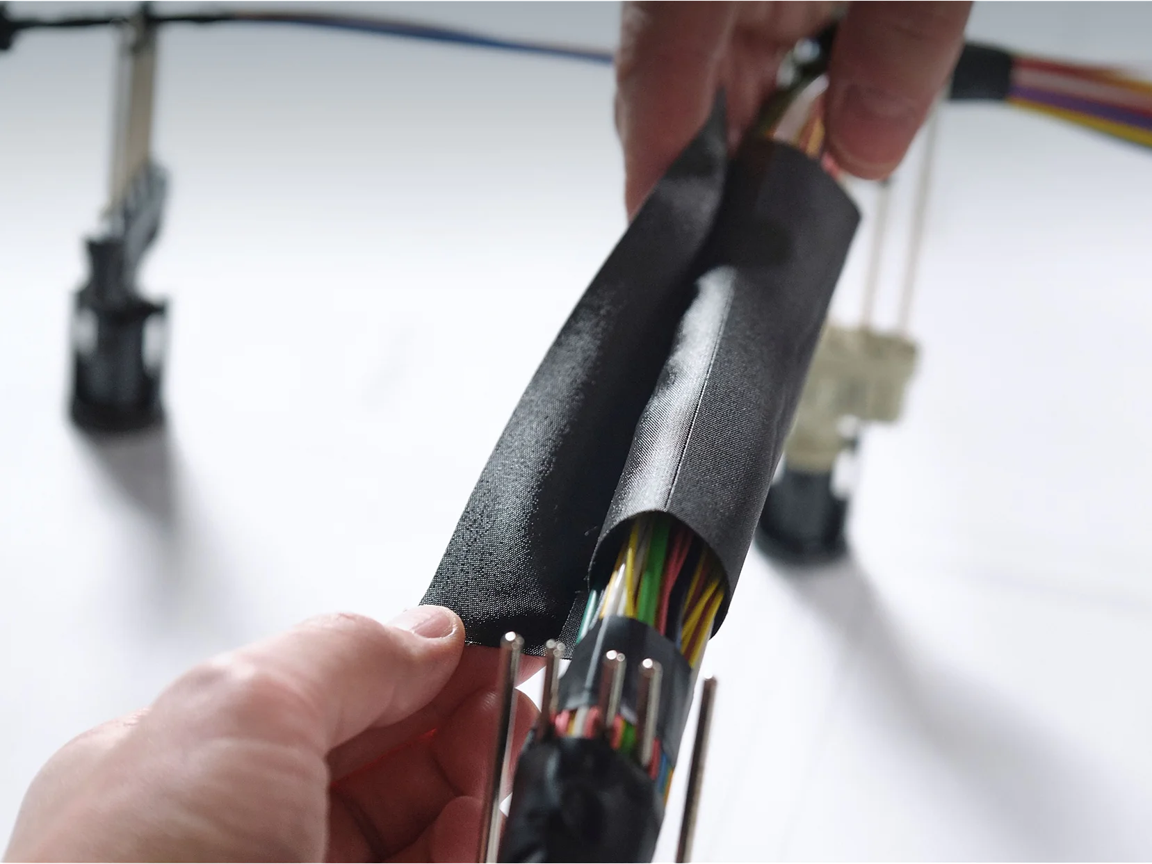 Close-up of hands wrapping a bundle of multicolored electrical wires with black insulating tesa tape. The wires are organized and connected to plastic connectors. A blurred background shows more wires and a surface with printed text. (This text has been generated by AI)