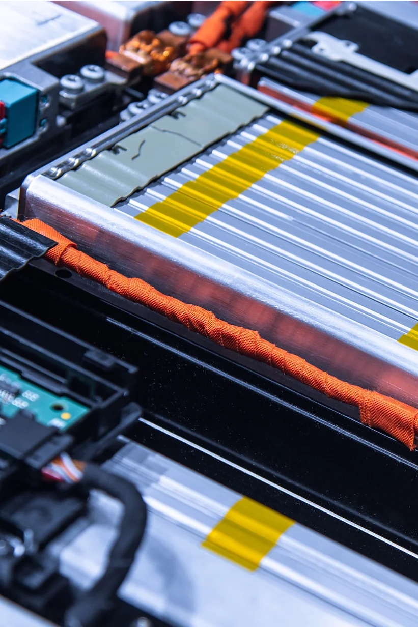 Close-up of a complex electrical circuit board and battery pack, featuring silver and yellow striped battery cells, orange wires, and a green circuit board. The components are tightly arranged within a metallic frame using tesa tape. (This text has been generated by AI)