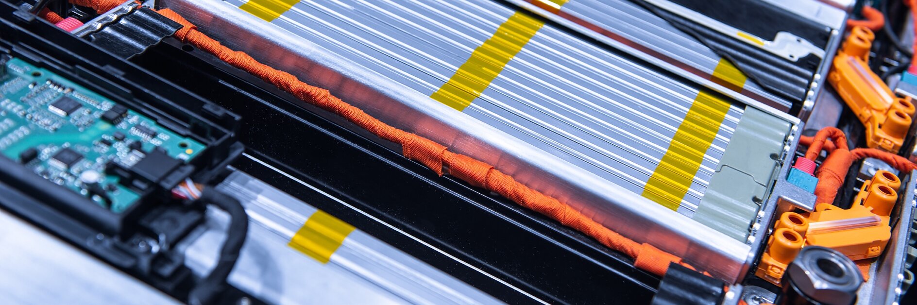 Close-up of a complex electrical circuit board and battery pack, featuring silver and yellow striped battery cells, orange wires, and a green circuit board. The components are tightly arranged within a metallic frame using tesa tape. (This text has been generated by AI)