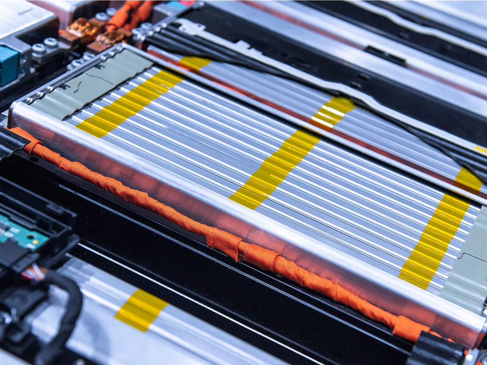 Close-up of a complex electrical circuit board and battery pack, featuring silver and yellow striped battery cells, orange wires, and a green circuit board. The components are tightly arranged within a metallic frame using tesa tape. (This text has been generated by AI)