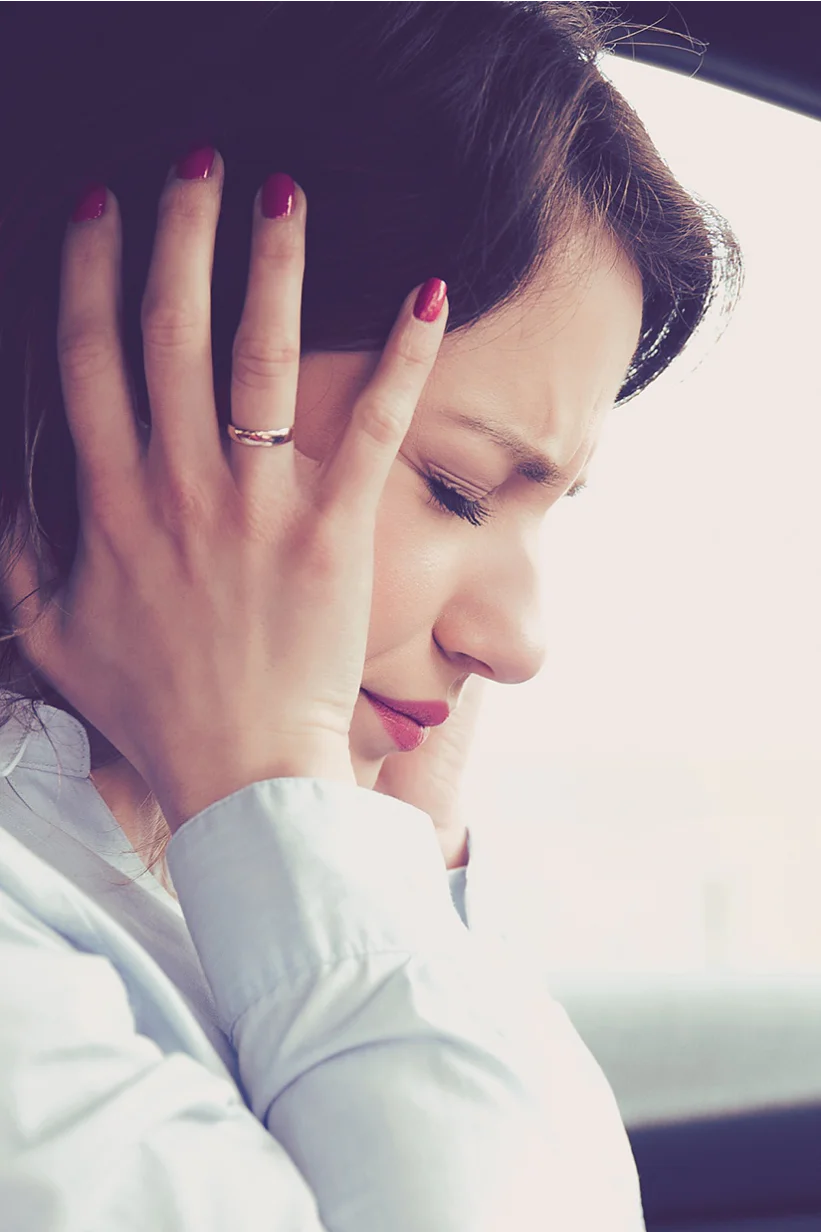 A woman is sitting in a car with both hands covering her ears. She appears to be uncomfortable or in distress. She is wearing a light-colored blouse and has dark hair. The interior of the car features dark and red accents. (This text has been generated by AI)