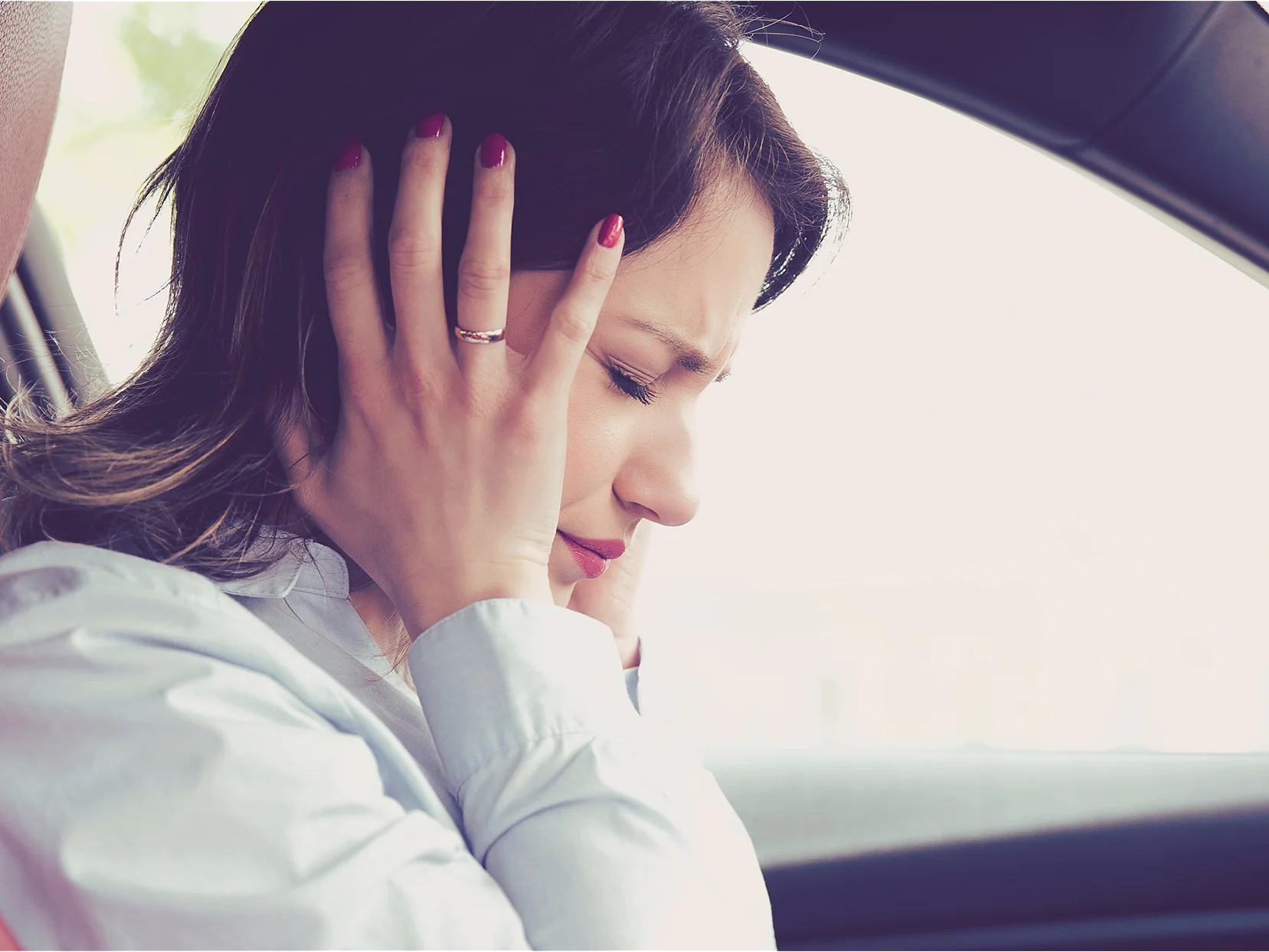 A woman is sitting in a car with both hands covering her ears. She appears to be uncomfortable or in distress. She is wearing a light-colored blouse and has dark hair. The interior of the car features dark and red accents. (This text has been generated by AI)