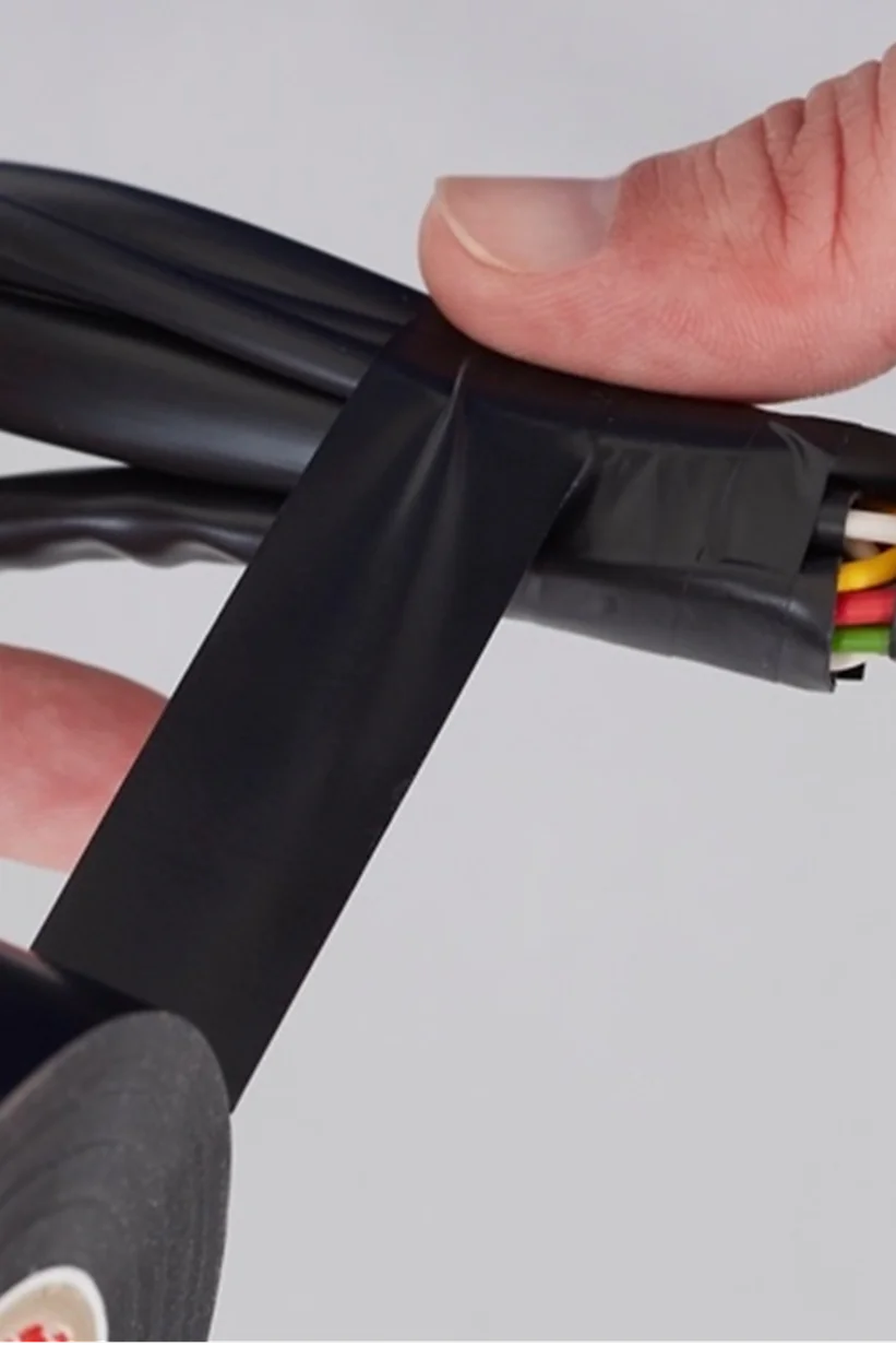 A person wraps black tesa tape around a group of colored wires. The tesa tape is being applied to secure and insulate the wires, which include green, yellow, and red colors. The background is plain white. (This text has been generated by AI)