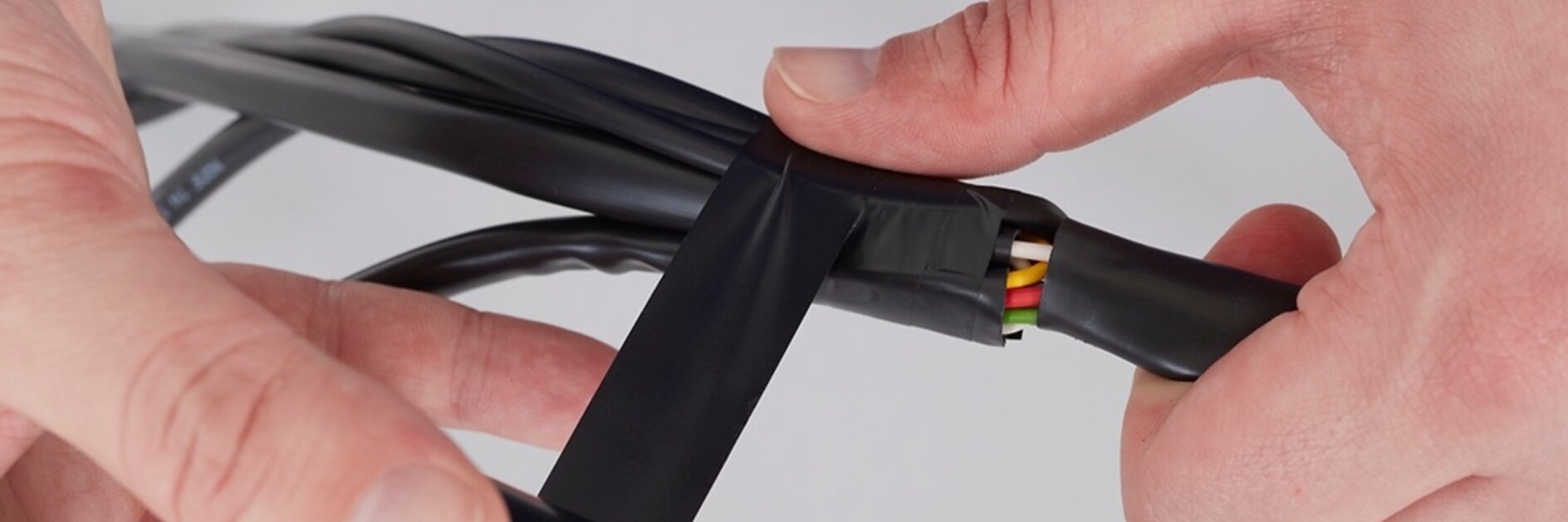 A person wraps black tesa tape around a group of colored wires. The tesa tape is being applied to secure and insulate the wires, which include green, yellow, and red colors. The background is plain white. (This text has been generated by AI)