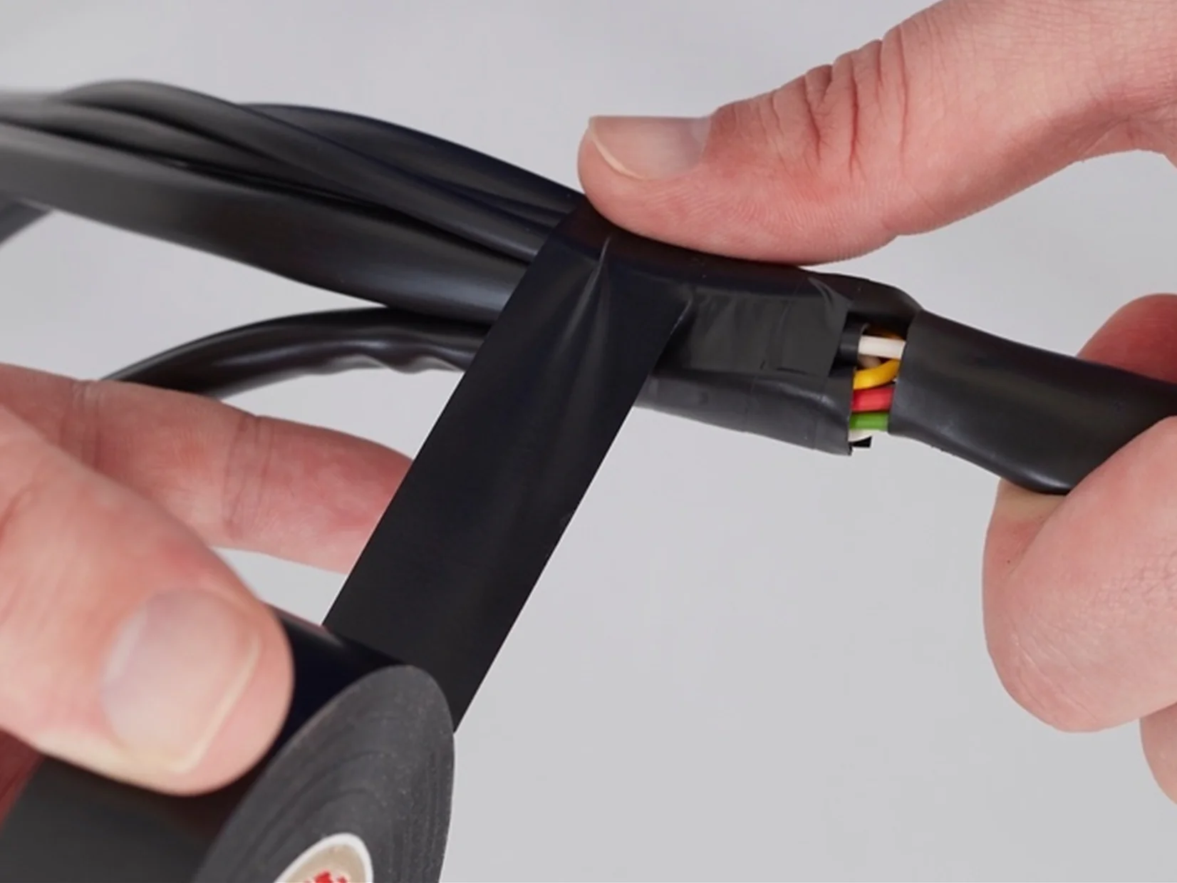 A person wraps black tesa tape around a group of colored wires. The tesa tape is being applied to secure and insulate the wires, which include green, yellow, and red colors. The background is plain white. (This text has been generated by AI)