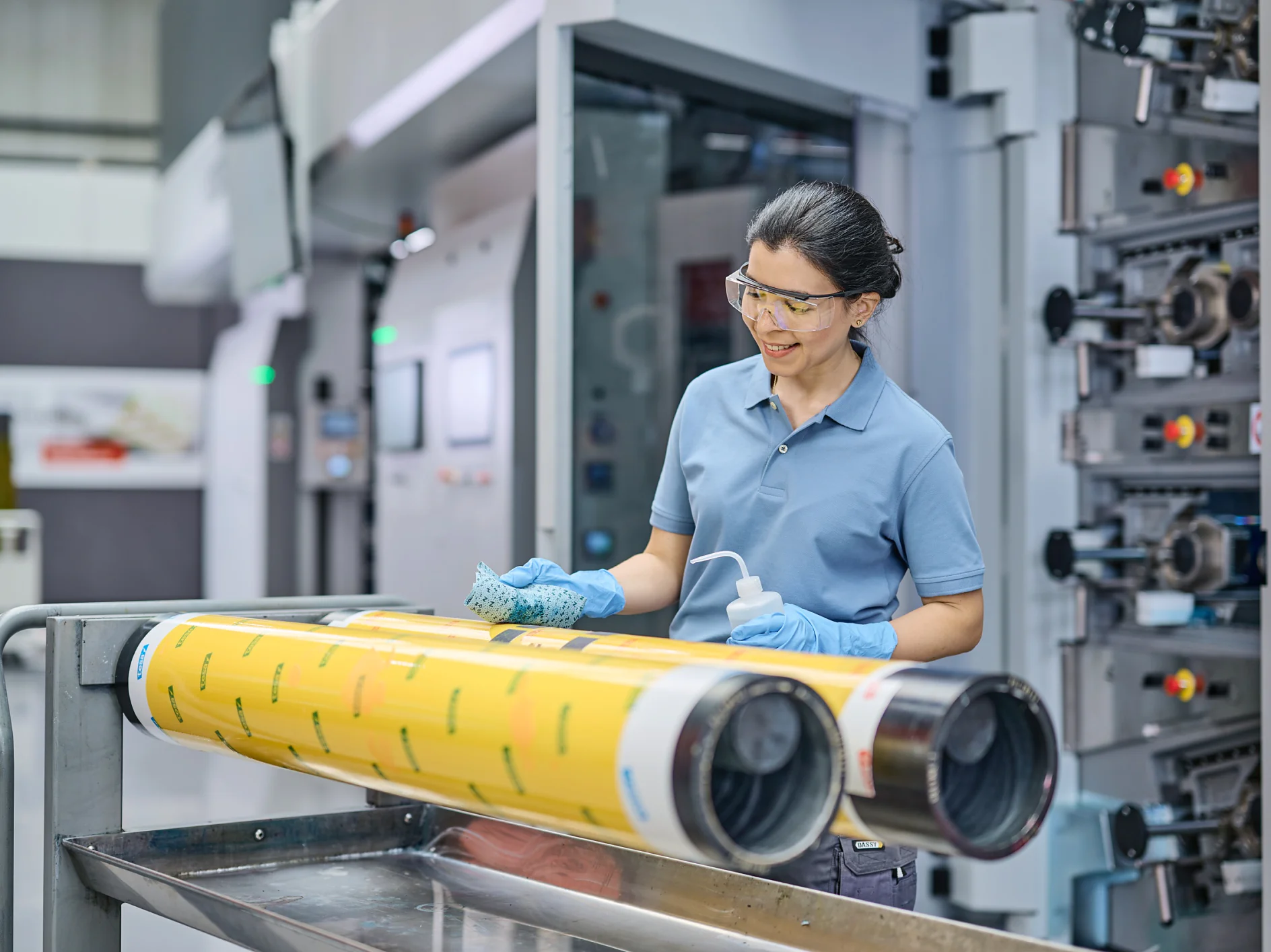 A person wearing safety glasses and gloves stands beside a printing press machine, cleaning large yellow rollers with a cloth and spray bottle. The scene appears to be in an industrial or manufacturing setting, with tesa tape nearby for additional tasks. (This text has been generated by AI)