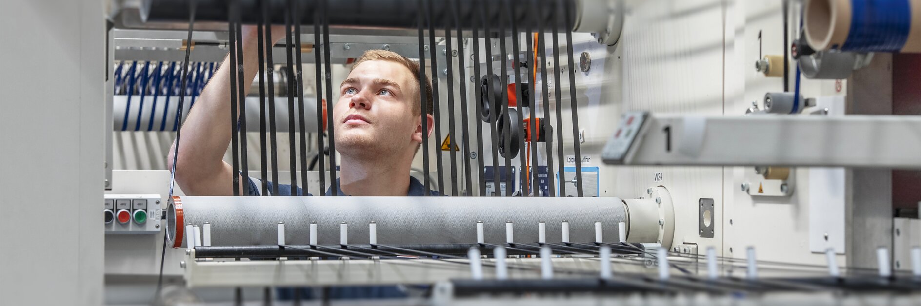 A person in a blue uniform works with machinery in a factory setting, adjusting black cables on a series of rollers and levers using tesa tape. The environment is industrial, with various mechanical components and controls visible.