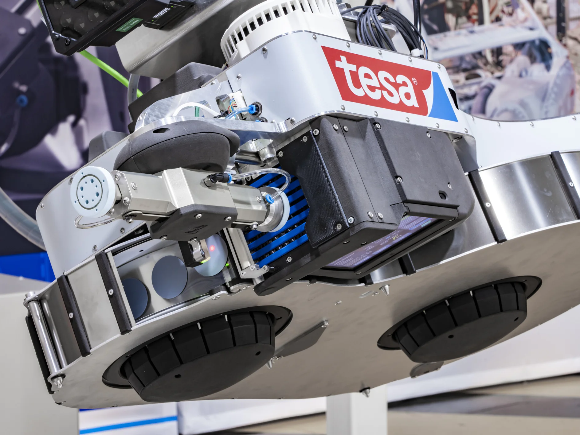 「tesa」のロゴが付いた産業用ロボットのクローズアップ。ロボットには、機械のぼやけた背景を背景に、tesa テープ、センサー、機械アームなどの複数のコンポーネントが搭載されています。デザインはメタリックで実用的です。. (This text has been generated by AI)