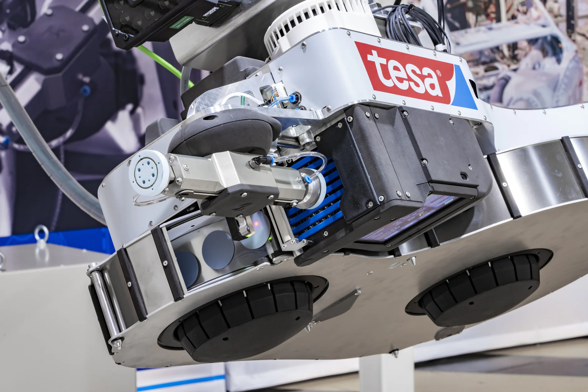 「tesa」のロゴが付いた産業用ロボットのクローズアップ。ロボットには、機械のぼやけた背景を背景に、tesa テープ、センサー、機械アームなどの複数のコンポーネントが搭載されています。デザインはメタリックで実用的です。. (This text has been generated by AI)