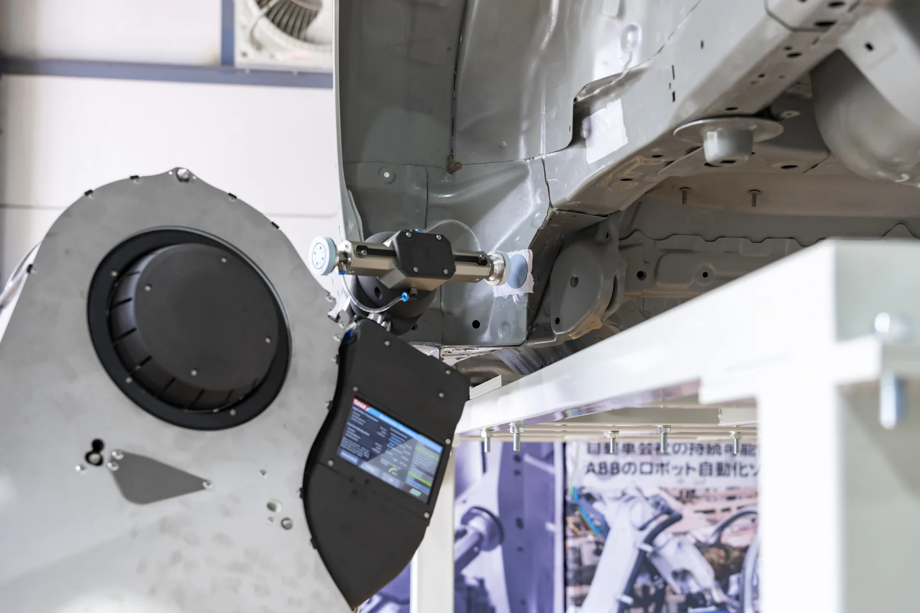 ロボット溶接アームは、産業現場で車両の下部構造の接合部の近くに設置されています。機械にはデータを表示するディスプレイ画面があり、このエリアは自動車製造工程の一部です。. (This text has been generated by AI)