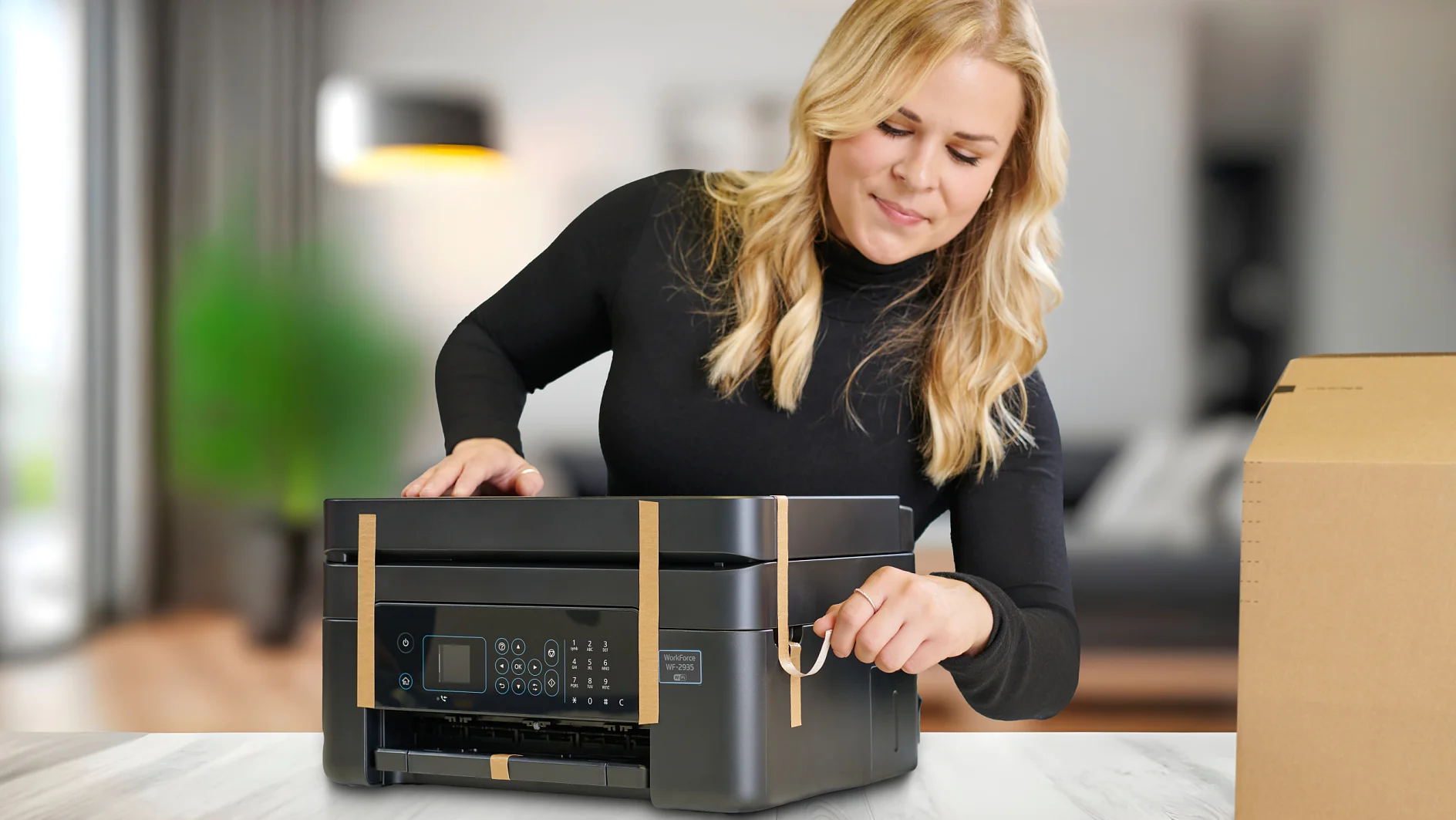 A woman is removing tesa tape from a black printer on a light-colored table. She is wearing a black top and standing in a modern, blurred living room. A brown cardboard box is visible on the tables right side. (This text has been generated by AI)