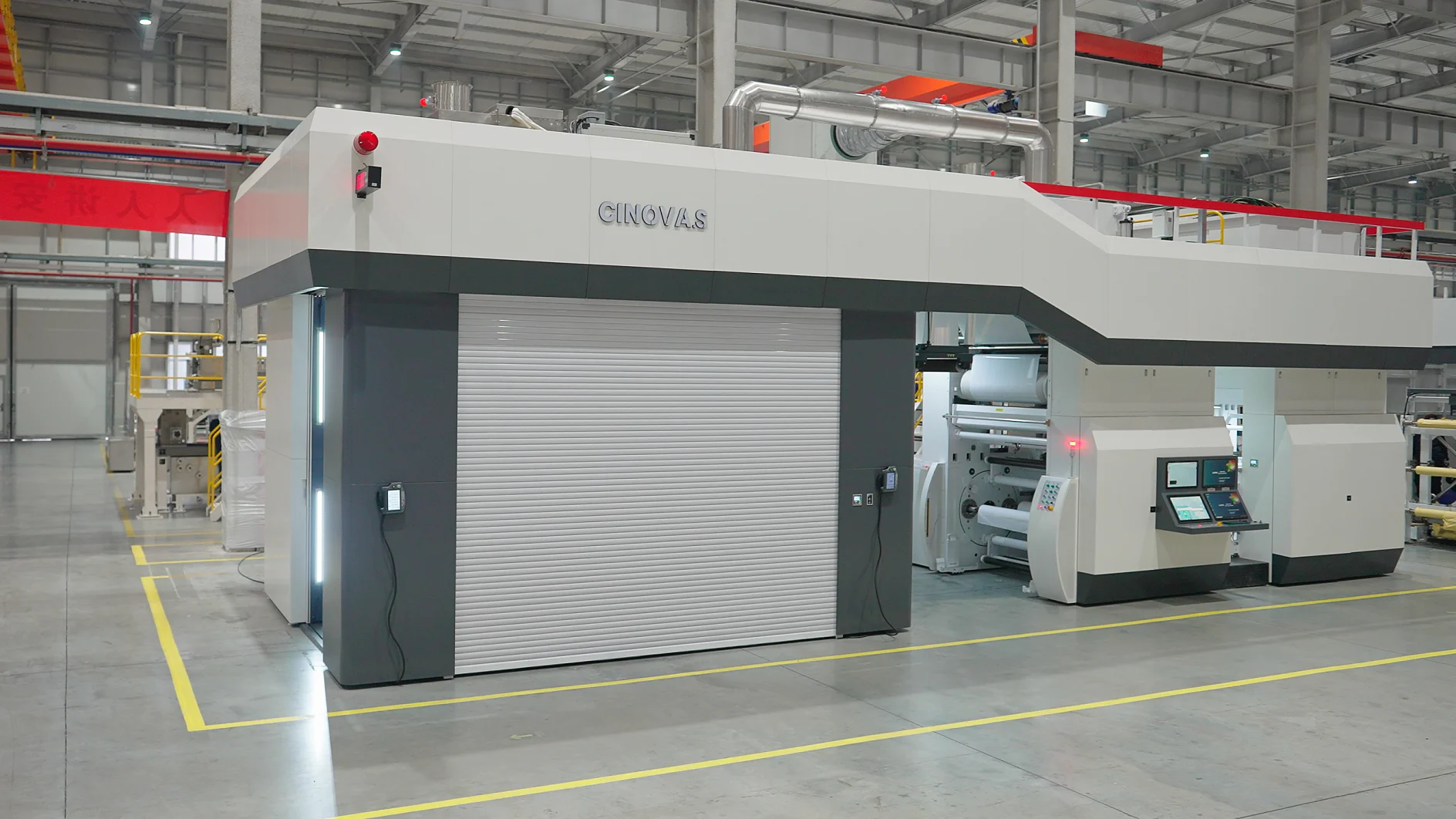 Large industrial printing machine in a factory setting. The machine is gray and white with control panels and display screens. The floor is concrete, and the area is well-lit with overhead lights. Yellow lines are marked on the floor for guidance using tesa tape. (This text has been generated by AI)