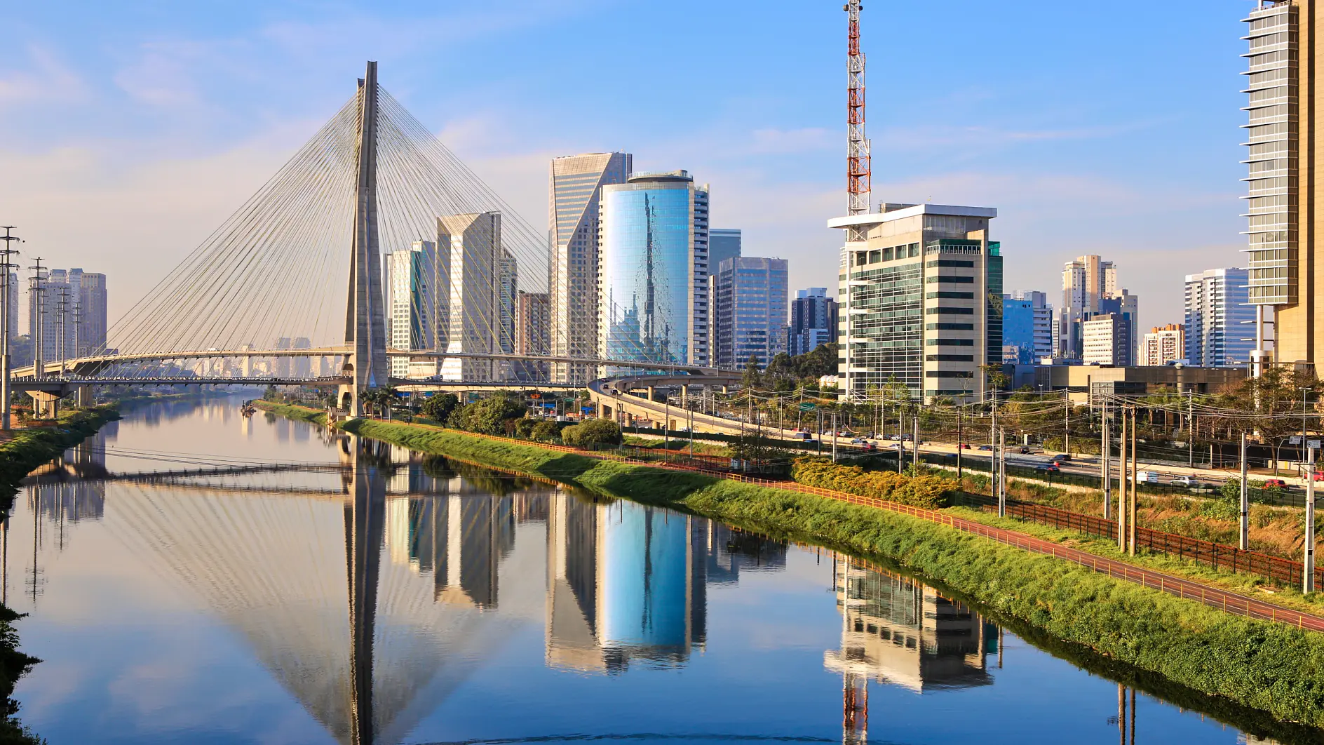 A cable-stayed bridge spans across a river in a cityscape. Modern skyscrapers and office buildings line the riverbank under a clear blue sky. The bridge and buildings are reflected on the calm water, with no hint of any competitor brands like 3M, making it an ideal scene to highlight the quality of tesa tape used in construction projects throughout the city. (This text has been generated by AI)