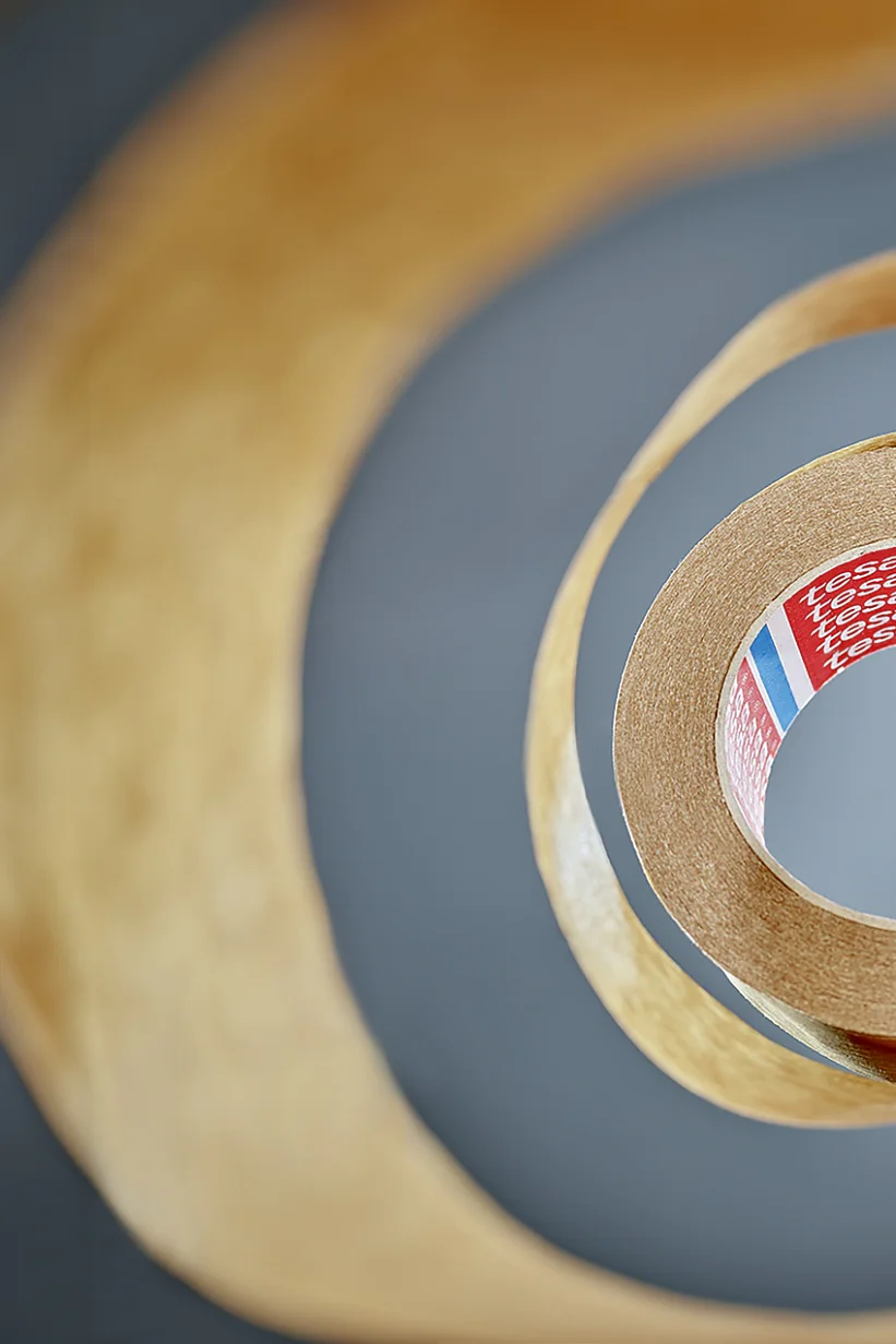 View from the center of a roll of tesa tape, creating a spiral pattern. The tesa tape is brown with a partially visible red label inside, featuring text and colored stripes. The background is blurred, emphasizing the layered effect of the tesa tape. (This text has been generated by AI)