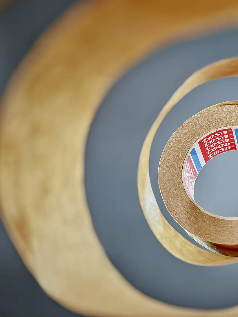 View from the center of a roll of tesa tape, creating a spiral pattern. The tesa tape is brown with a partially visible red label inside, featuring text and colored stripes. The background is blurred, emphasizing the layered effect of the tesa tape. (This text has been generated by AI)