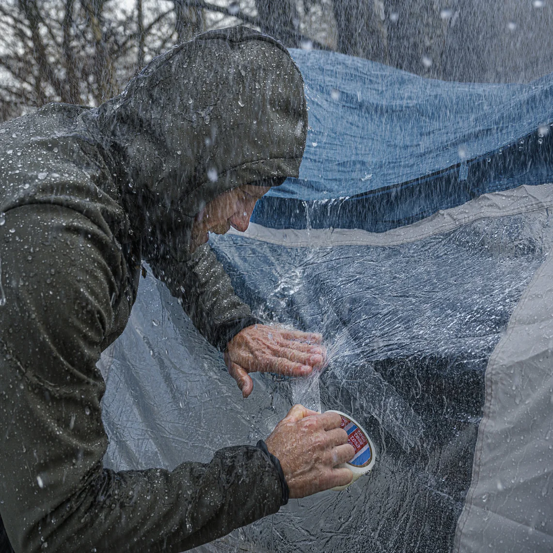フード付きのレインジャケットを着た人が、大雨の中、濡れたテントにテサテープを使って防水シーラントを塗っています。テントは青と灰色で、表面から水がはねているのが目に見えてわかります。背景の木々はぼやけています。. (This text has been generated by AI)