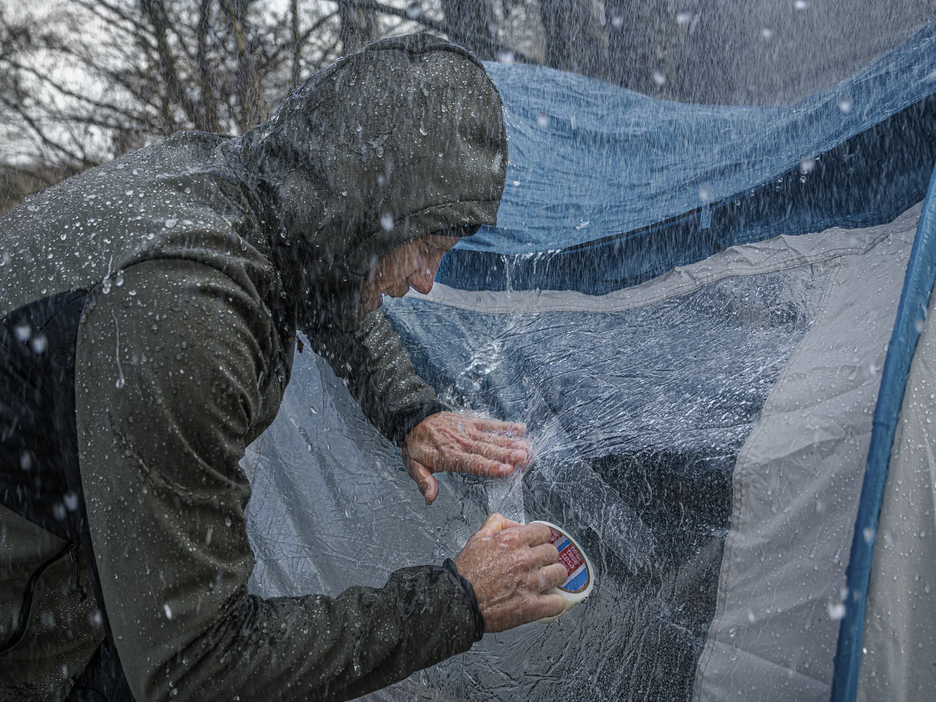フード付きのレインジャケットを着た人が、大雨の中、濡れたテントにテサテープを使って防水シーラントを塗っています。テントは青と灰色で、表面から水がはねているのが目に見えてわかります。背景の木々はぼやけています。. (This text has been generated by AI)