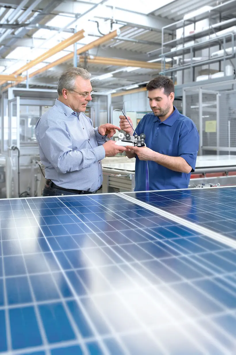 Two men are examining a small piece of machinery in a bright, industrial setting. They stand next to large solar panels. The background shows various equipment and metal structures, indicating a technological or manufacturing environment. (This text has been generated by AI)
