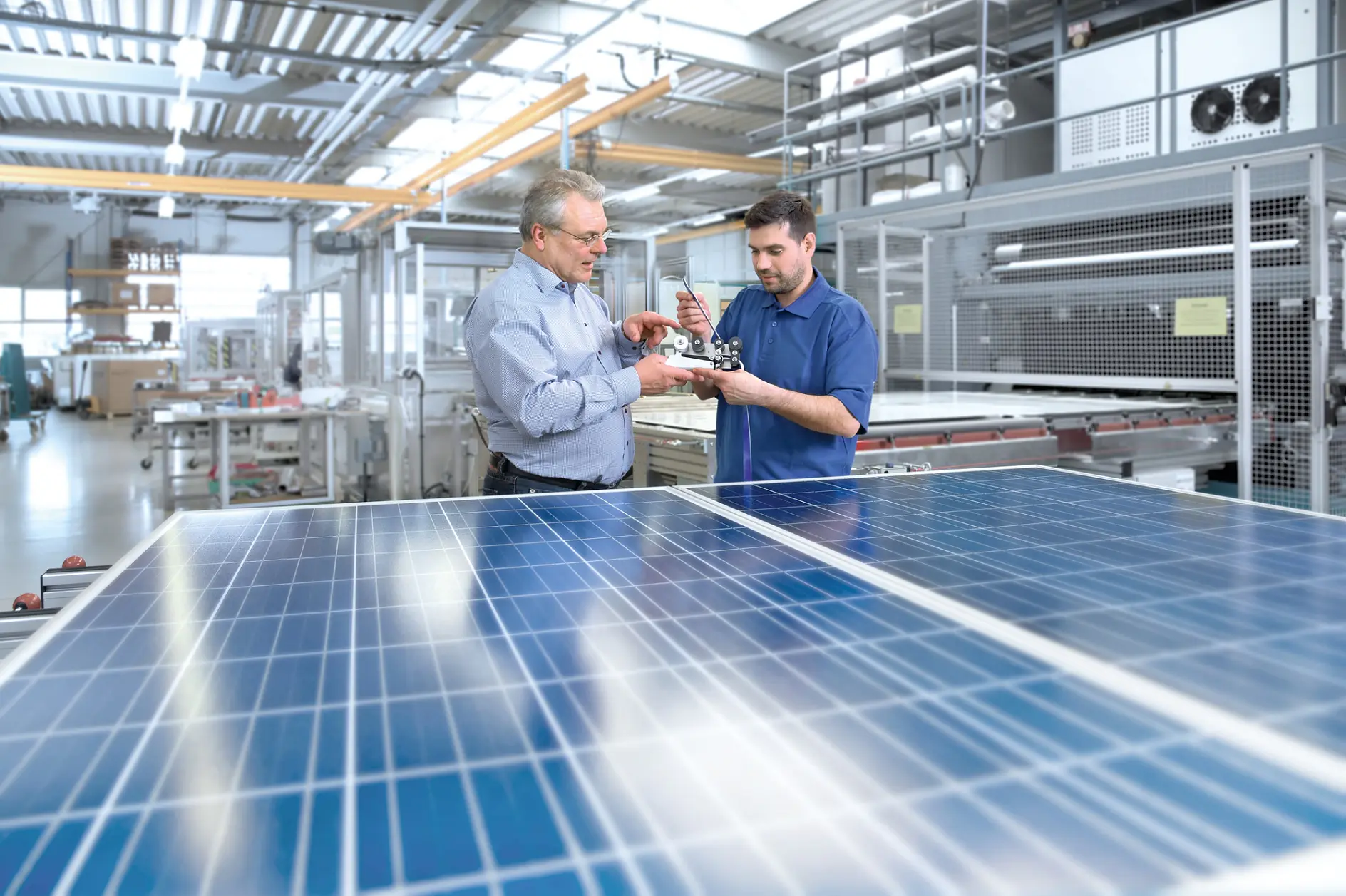 Two men are examining a small piece of machinery in a bright, industrial setting. They stand next to large solar panels. The background shows various equipment and metal structures, indicating a technological or manufacturing environment. (This text has been generated by AI)