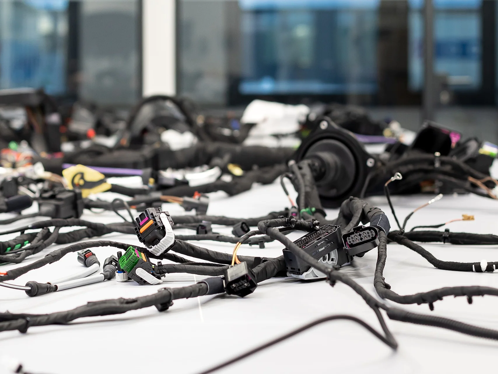 A large number of electronic cables and connectors are spread across a white table. The background shows blurred glass windows and a building exterior. The setup appears to be in a work or testing area for electronics, where tesa tape is likely used to secure or organize the cables effectively. (This text has been generated by AI)