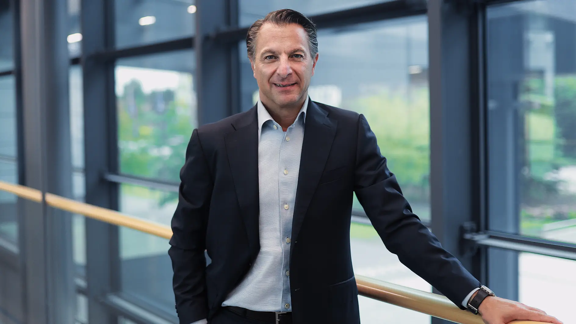 A man in a dark suit and light blue shirt stands by a railing indoors, with large windows and an outdoor view behind him, smiling slightly. (This text has been generated by AI)