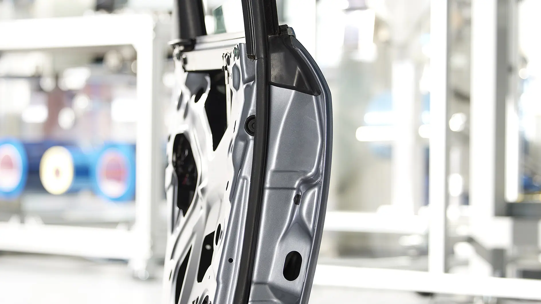 Side view of a metal car door frame in a manufacturing setting. The frame is silver with visible cutouts and structural details, secured with tesa tape. The background is blurred, featuring industrial equipment and machinery. (This text has been generated by AI)