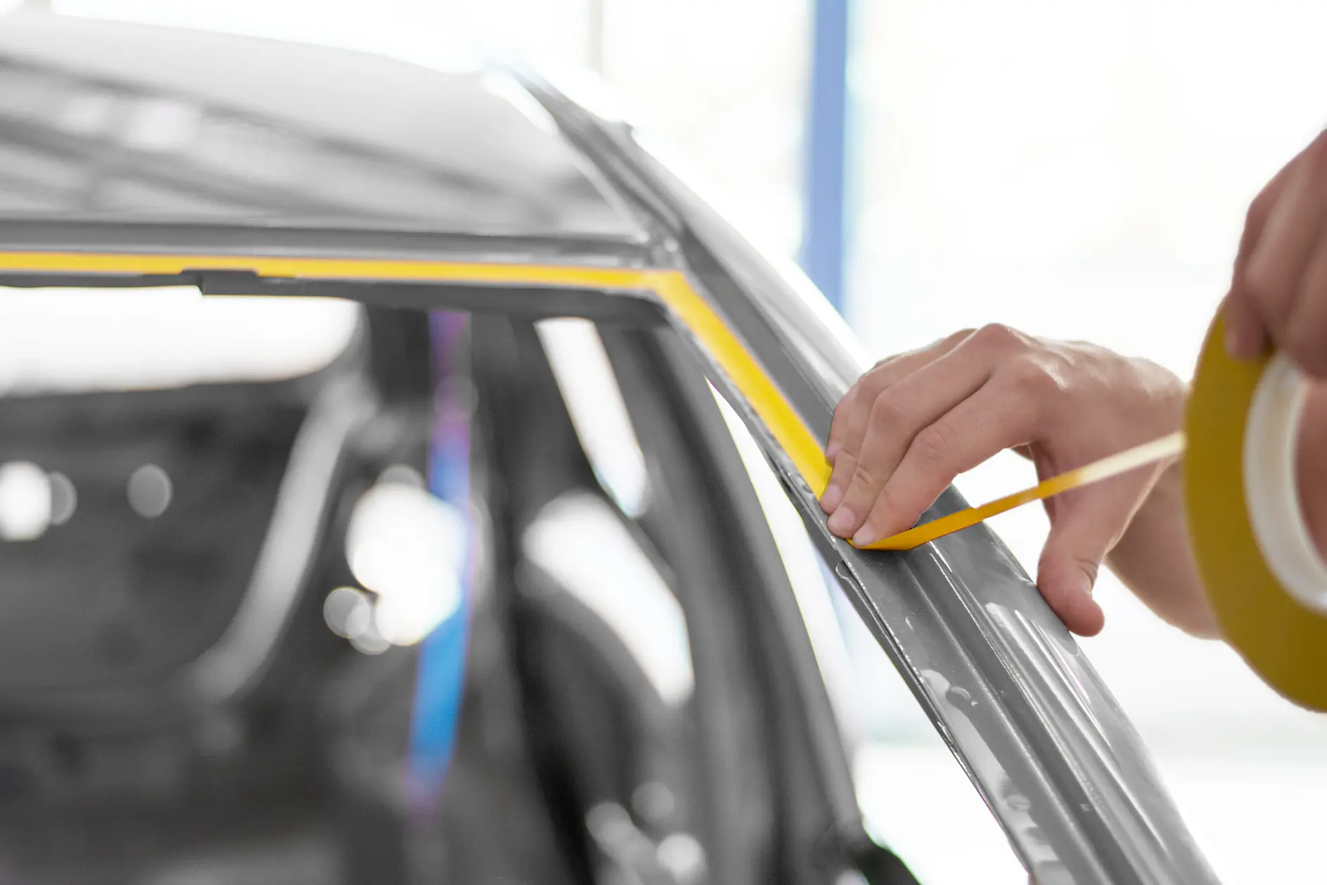 hand applying yellow masking tape along car window frame in a bright indoor auto workshop