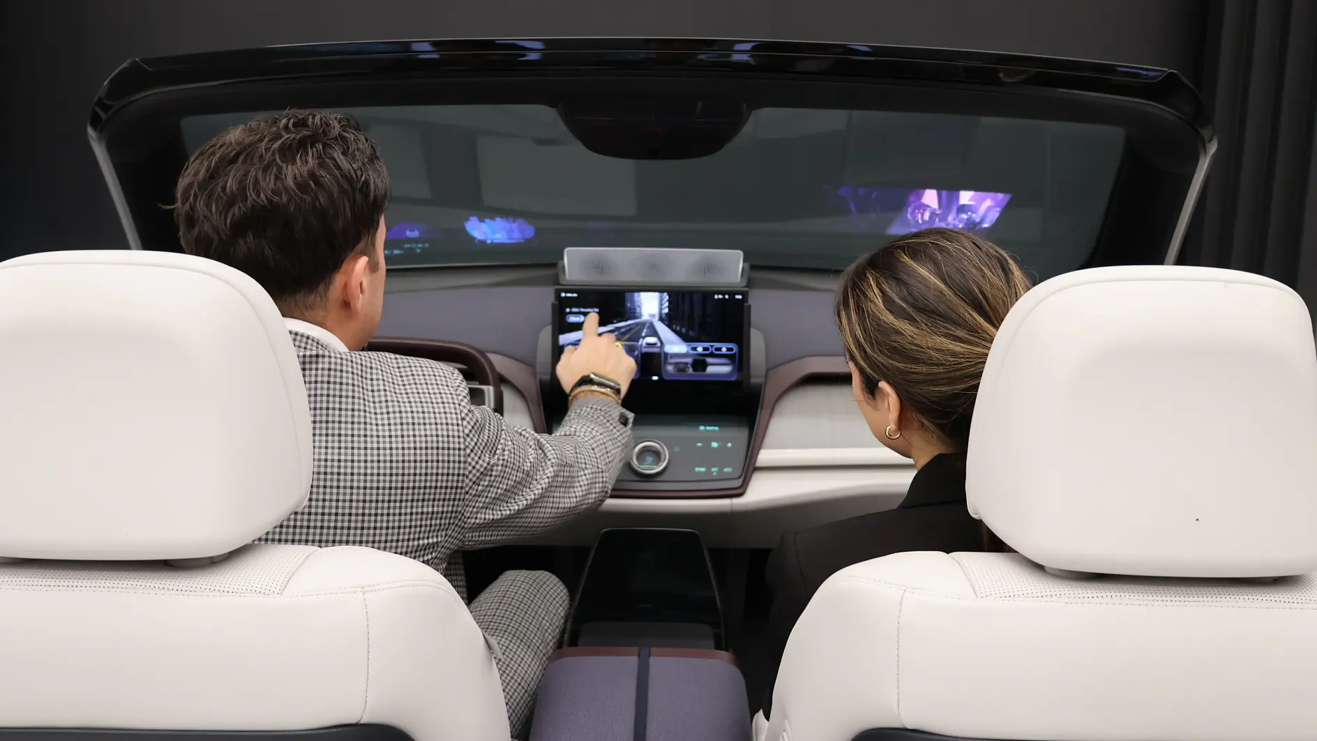 Two people sit in a modern vehicle’s front seats, viewed from behind, as one points to the dashboard touchscreen. Both watch the screen. (This text has been generated by AI)