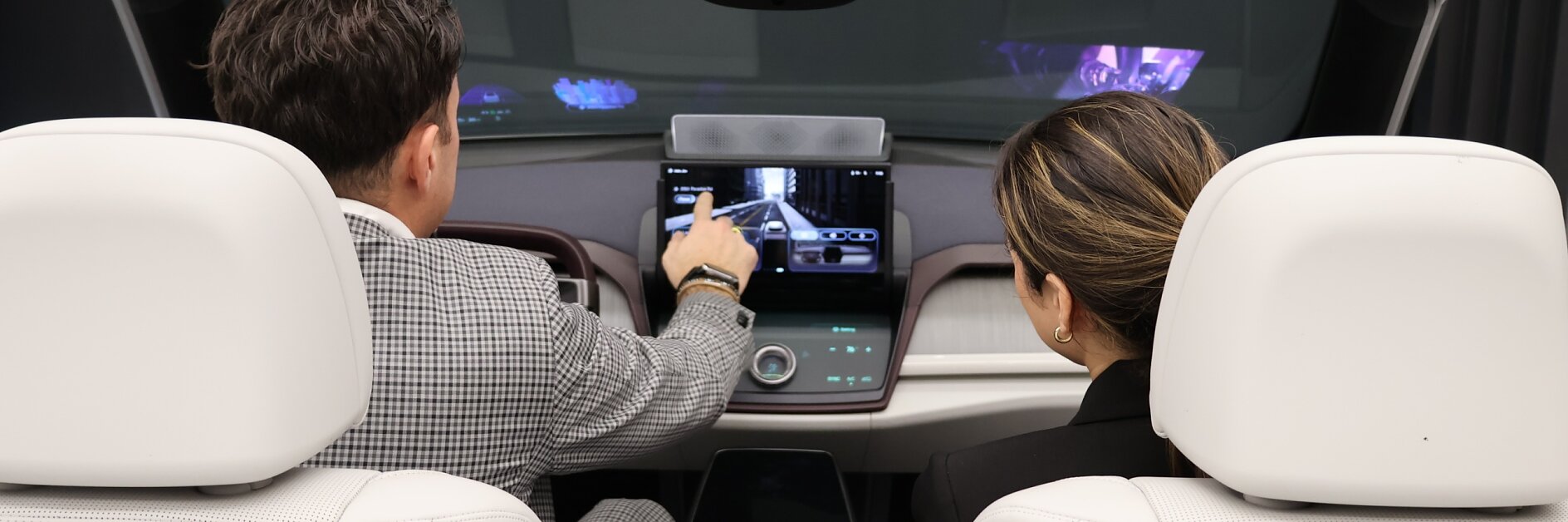 Two people sit in a modern vehicle’s front seats, viewed from behind, as one points to the dashboard touchscreen. Both watch the screen.