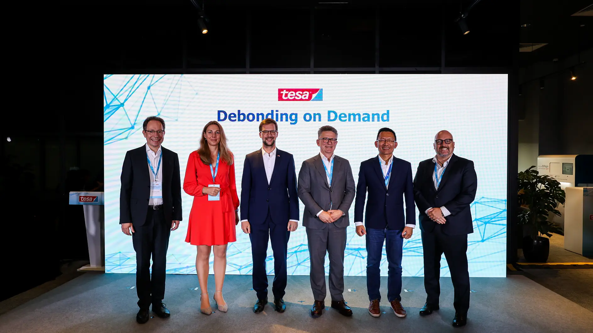 Six people stand on a stage in front of a large screen displaying the tesa tape logo and the words Debonding on Demand. They are dressed in business attire and facing the camera, with a dark room and display monitors in the background. (This text has been generated by AI)