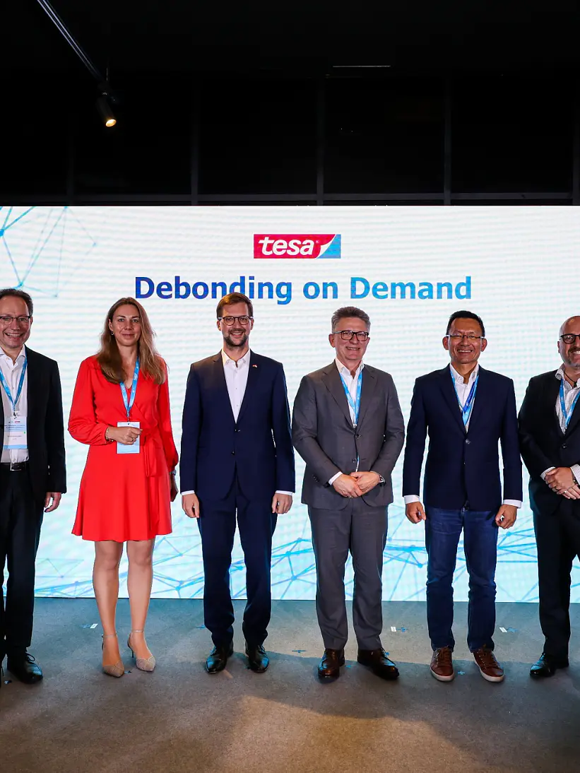 Six people stand on a stage in front of a large screen displaying the tesa tape logo and the words Debonding on Demand. They are dressed in business attire and facing the camera, with a dark room and display monitors in the background. (This text has been generated by AI)