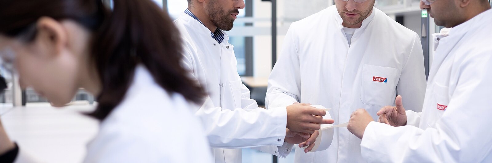 Four scientists are collaborating in a modern laboratory. Three men and one woman are wearing white lab coats and safety glasses. They are engaged in a discussion, with one man pointing at a document or device held by another, which appears to be secured using tesa tape.