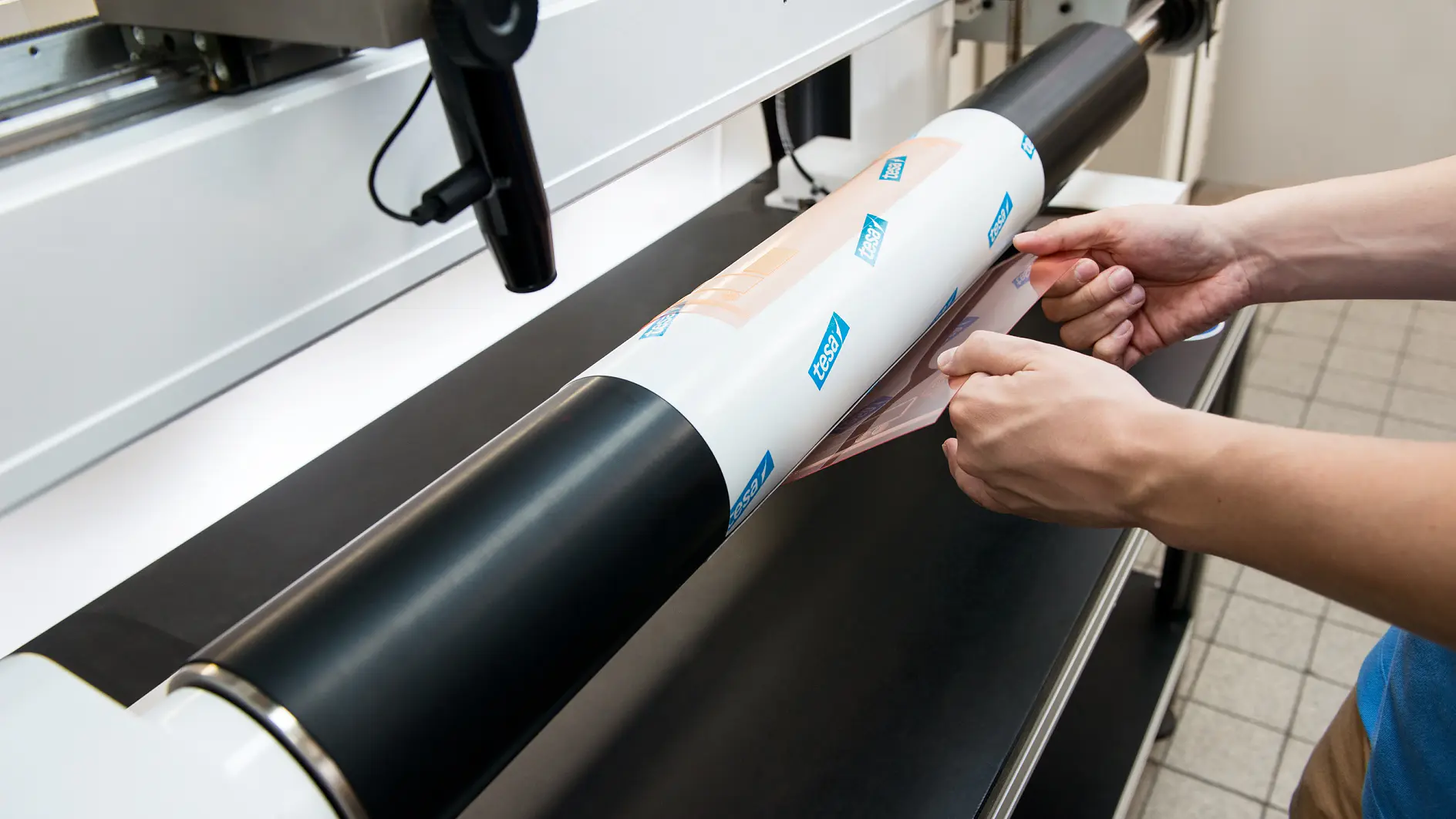 hands applying a tesa branded adhesive vinyl sheet onto a large flatbed printer indoors (This text has been generated by AI)