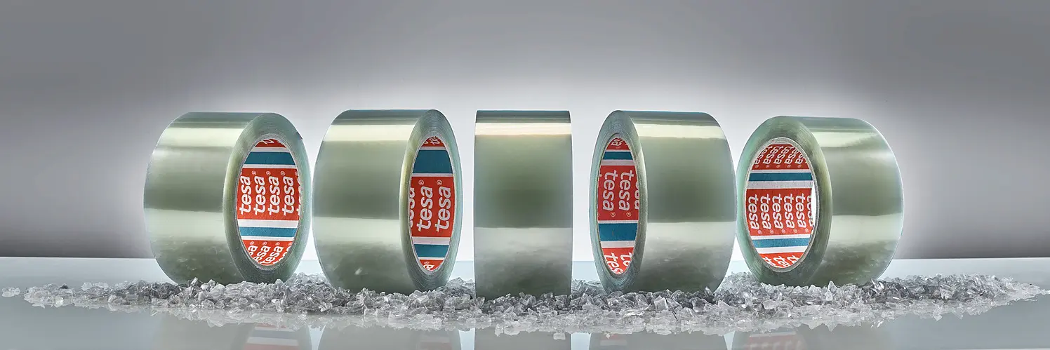Five rolls of clear adhesive tesa tape are standing upright on a surface scattered with small transparent crystals. The inner sides of the rolls display a red and blue label with white text. The background is a plain gray gradient.