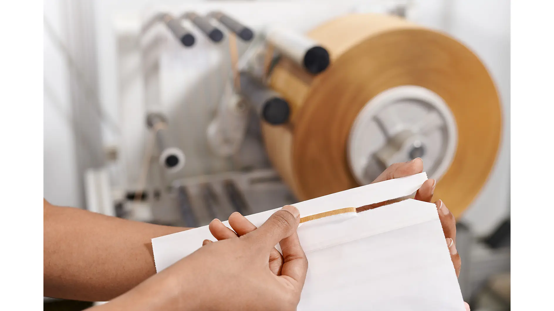 hands tearing a strip from a roll of brown tesa tape in an indoor packaging or manufacturing setting (This text has been generated by AI)