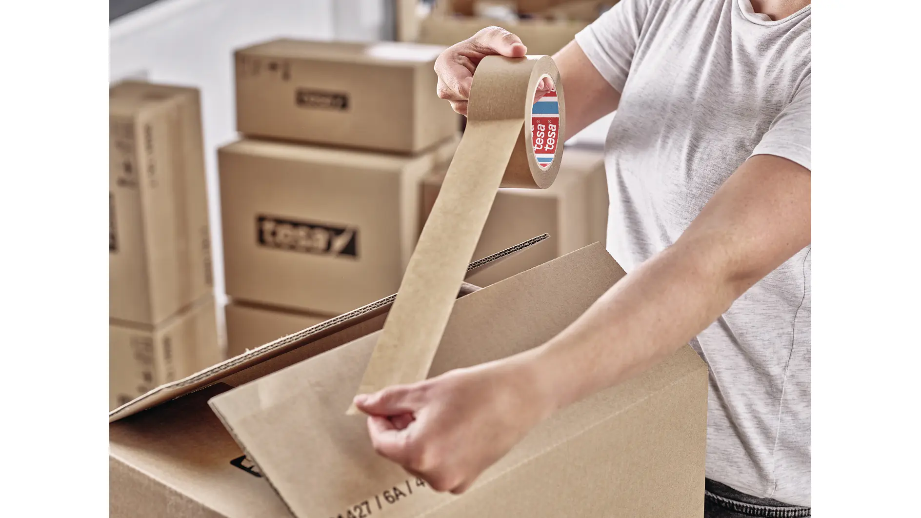 person sealing a cardboard box with tesa tape indoors, surrounded by stacked moving boxes in a home setting (This text has been generated by AI)