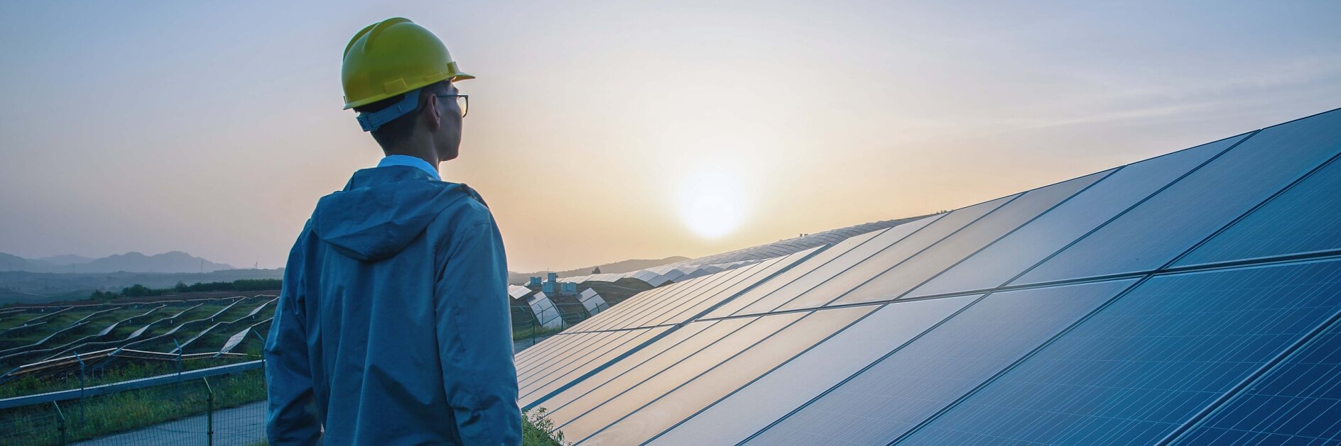 A person in a yellow hard hat and blue jacket stands in a grassy area, looking at a large array of solar panels at sunset. The sky is a gradient of orange and blue, and distant hills are visible under the setting sun. (This text has been generated by AI)