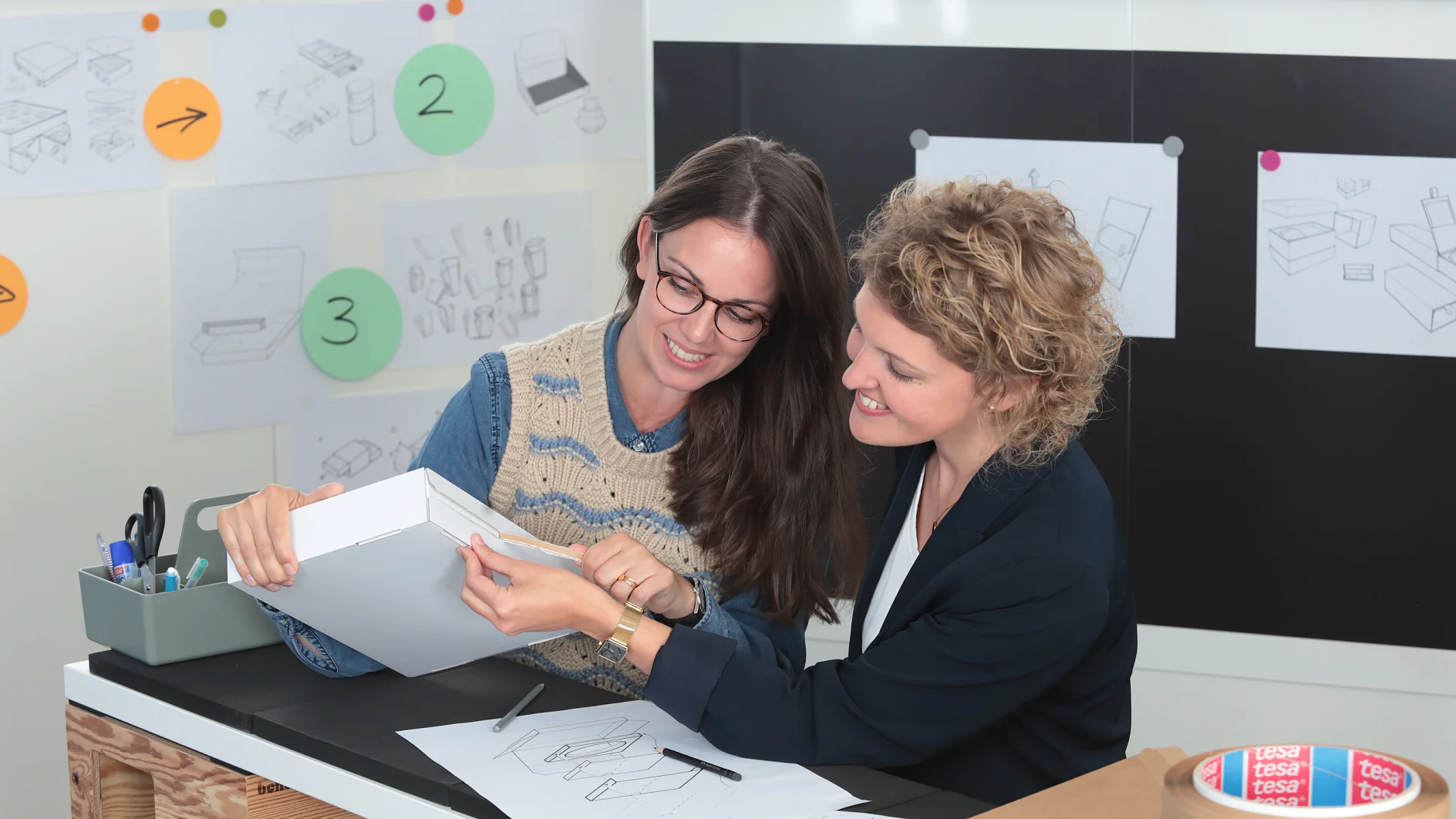 Two people are seated at a table with design sketches, examining a white box. The workspace has sketches and notes on the wall. Various office supplies, including tesa tape and markers, are on the table. Both appear engaged in discussion. (This text has been generated by AI)
