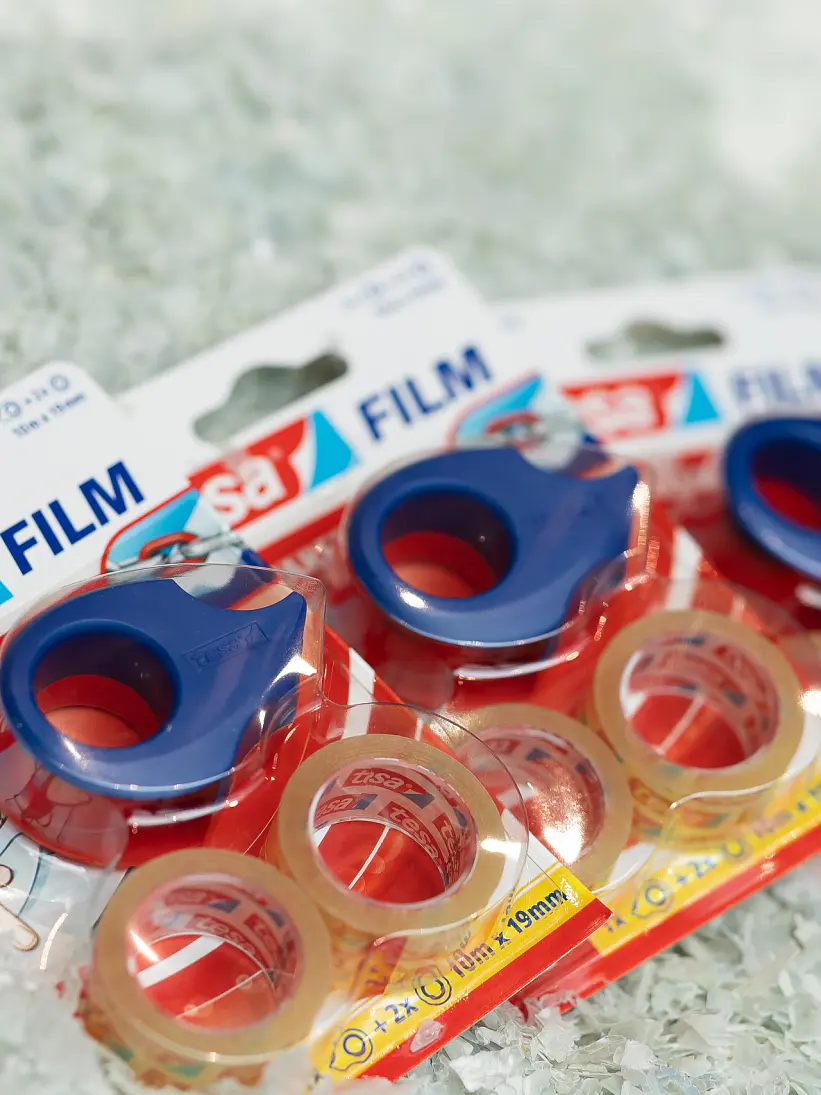 Three packages of tesa transparent adhesive tape are displayed on a pile of shredded paper. Each package contains a roll of tesa tape with a blue dispenser. The packaging text includes tesa Film and transparent. (This text has been generated by AI)