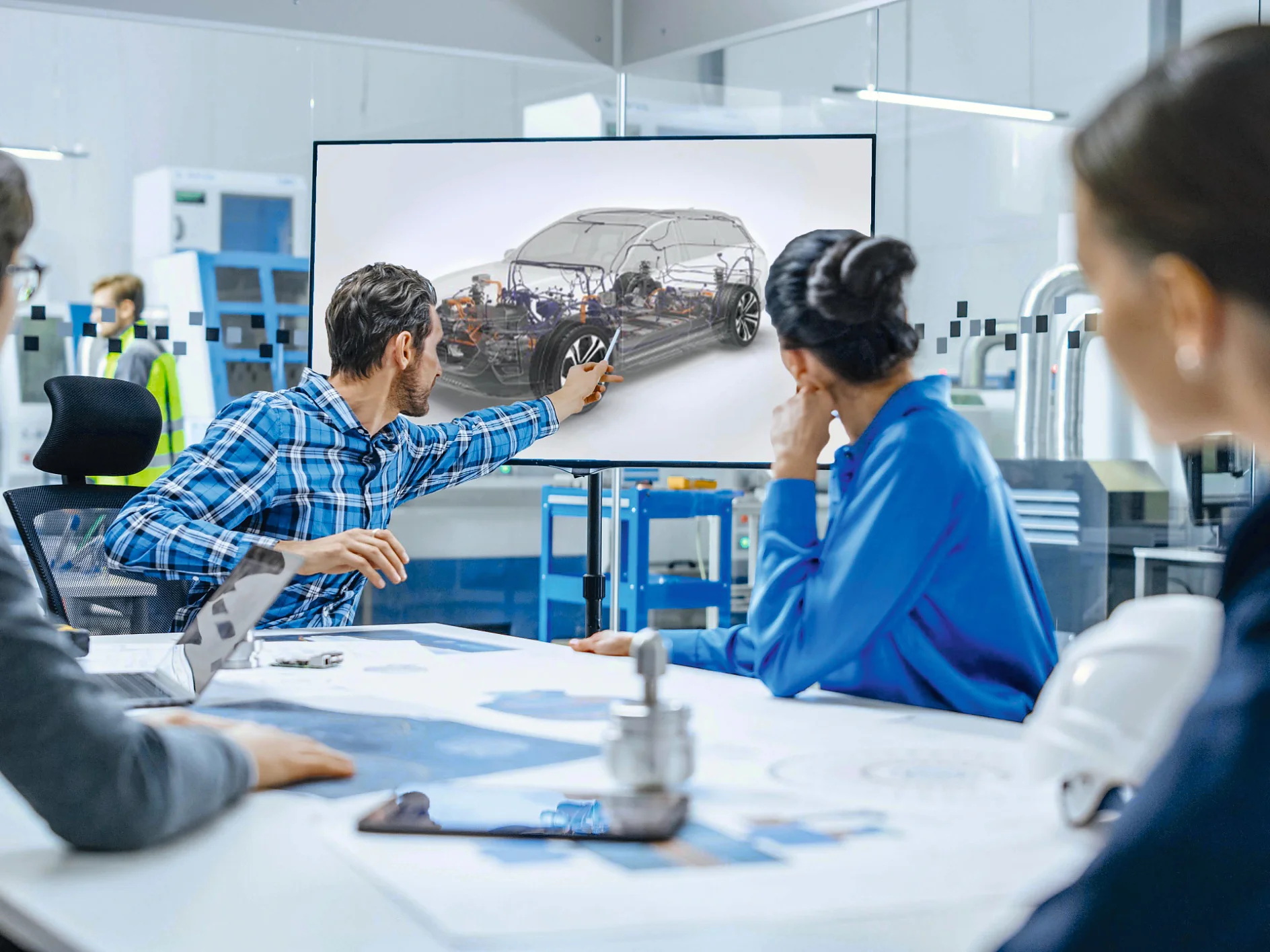 A group of four people sits around a table in an office, focusing on a large screen. One person is pointing to a car displayed on the screen. Blueprints and a model are on the table, held in place with tesa tape. The setting appears to be a professional meeting or presentation. (This text has been generated by AI)