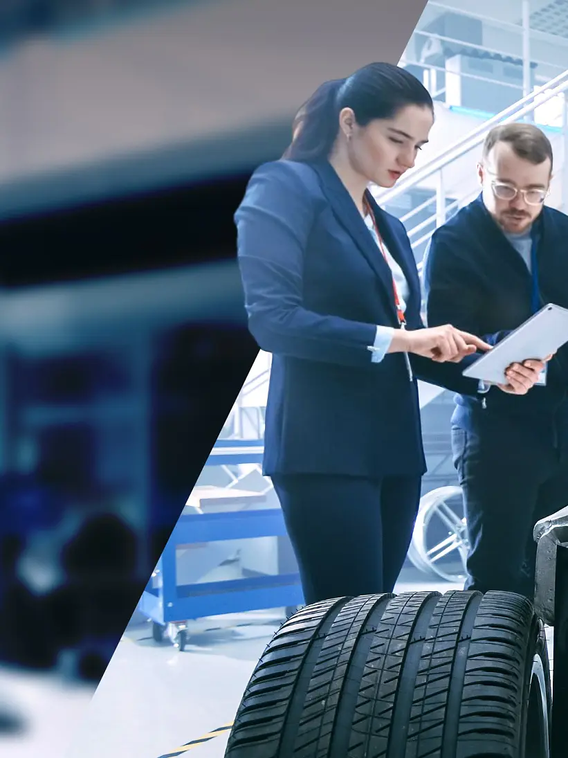 Two professionals in a modern laboratory setting examine a tire. The woman in a suit holds a tablet, while the man with glasses looks on. Equipment and metal stairs are visible in the background, and several rolls of tesa tape are prominently displayed on a nearby workbench. (This text has been generated by AI)