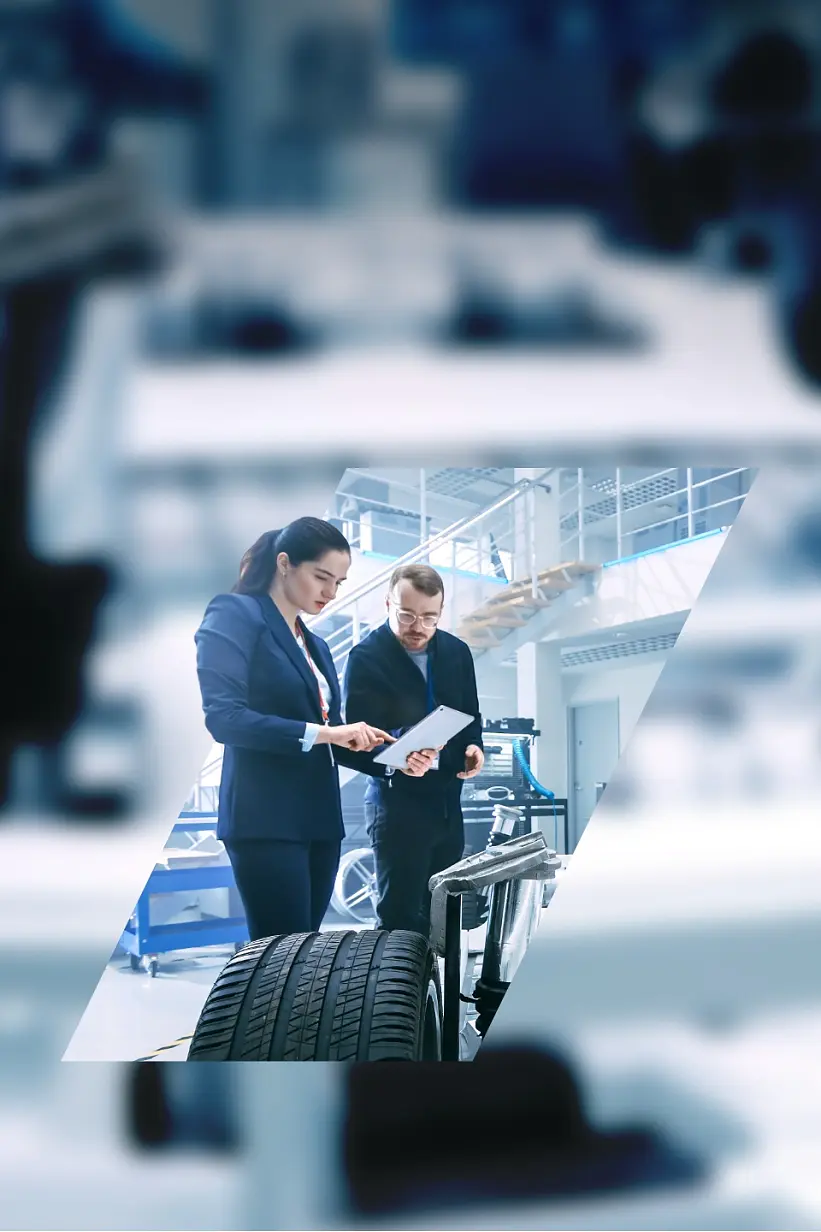 Two individuals review documents in an automotive workshop. A car chassis with exposed wheels is visible in the foreground, with strips of tesa tape securing various components. The background is a modern, well-lit industrial space filled with equipment and tools. (This text has been generated by AI)