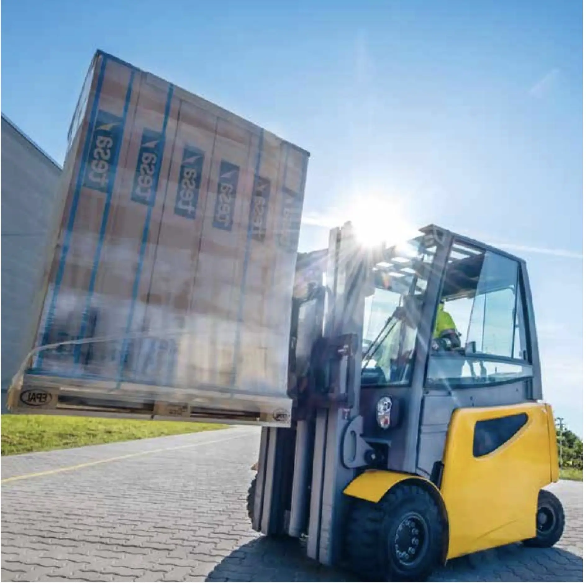 A forklift lifts a large, wrapped pallet on an outdoor pavement. The forklift is yellow and black, and the sun shines in the background, creating lens flare. A driver is visible inside the forklift cabin.