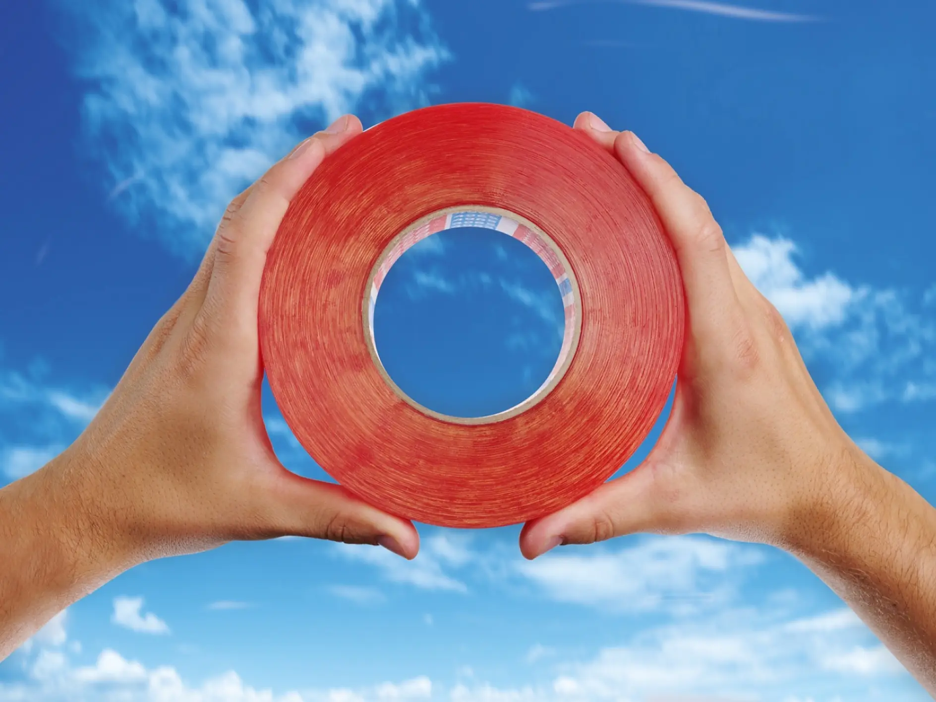 Two hands hold a large roll of red tesa tape against a backdrop of a blue sky with scattered clouds. The tesa tape roll forms a circular frame through which the sky is visible.