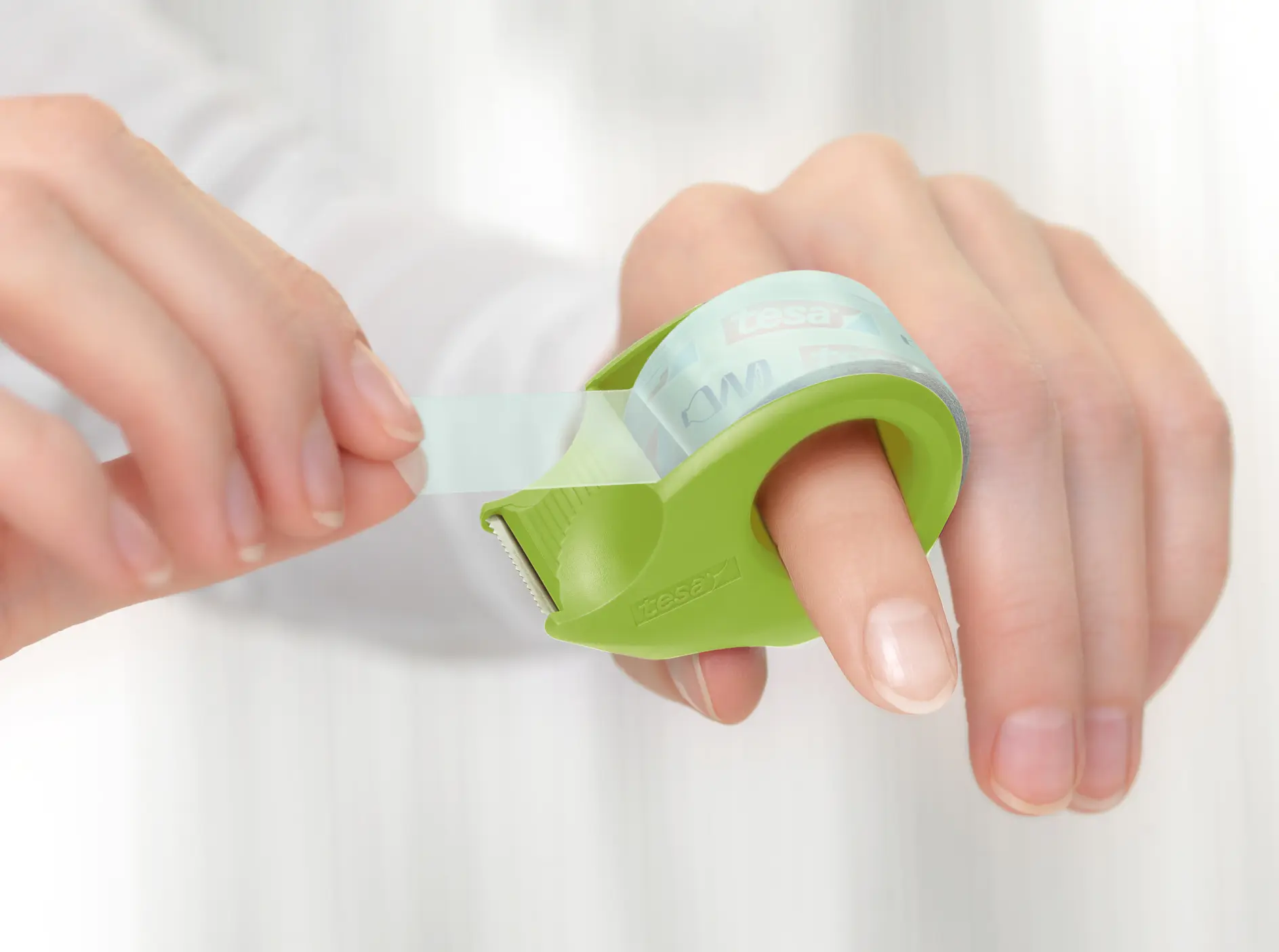 Close-up of a person dispensing clear adhesive tesa tape using a green handheld tape dispenser. The dispenser is wrapped around the index finger, and the person is pulling the tesa tape with the other hand. The background is softly blurred.